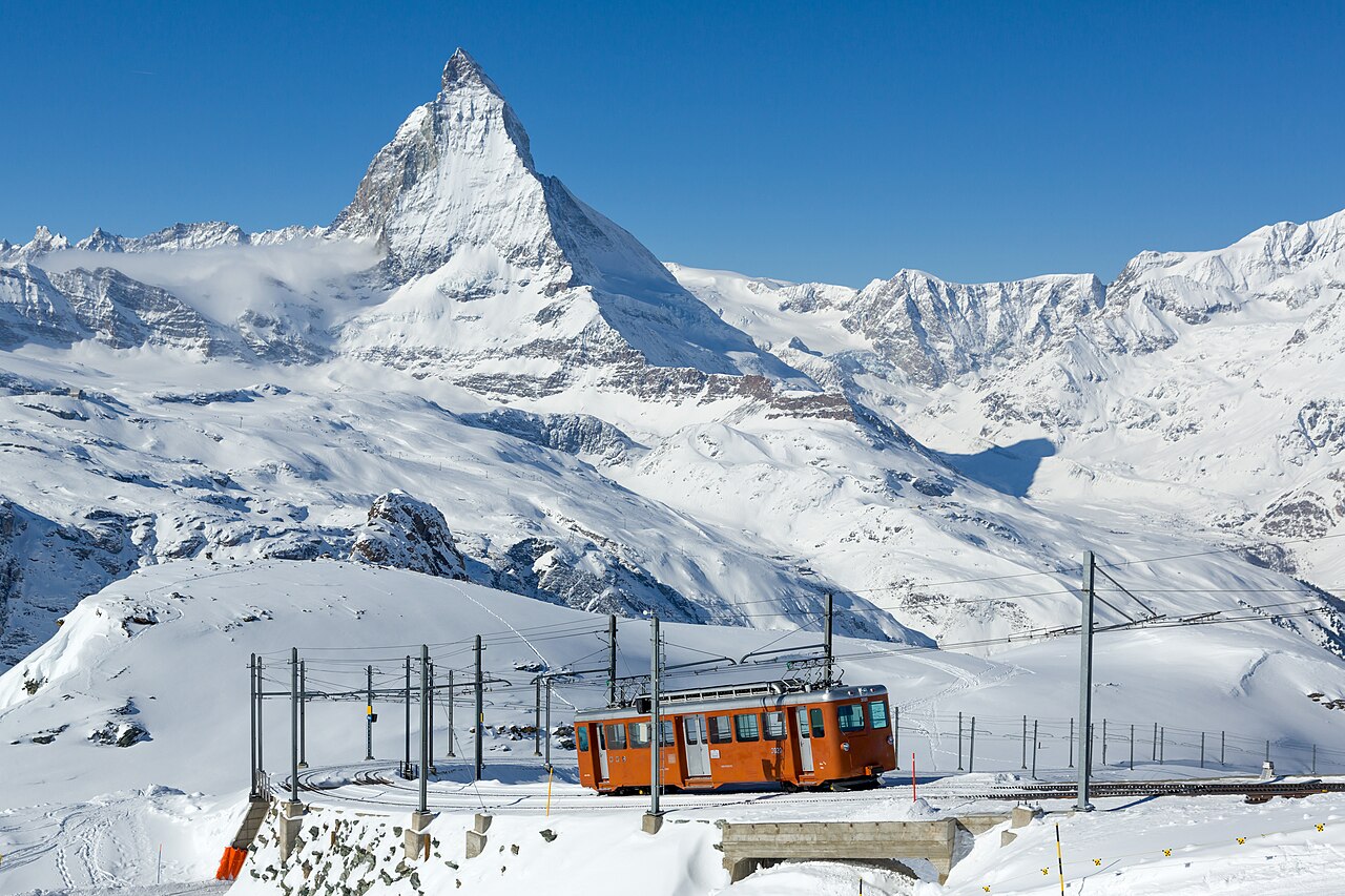 A short red mountain railway railcar on a snowy track, with a sharp rock peak rising behind it