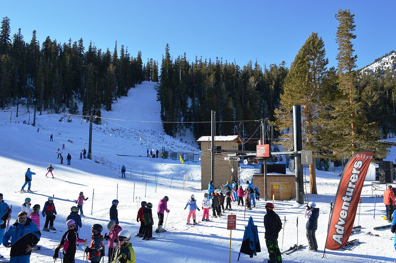 Skiers gathered on packed snow beside a surface lift outside a timber lodge, with pine trees on the slope behind