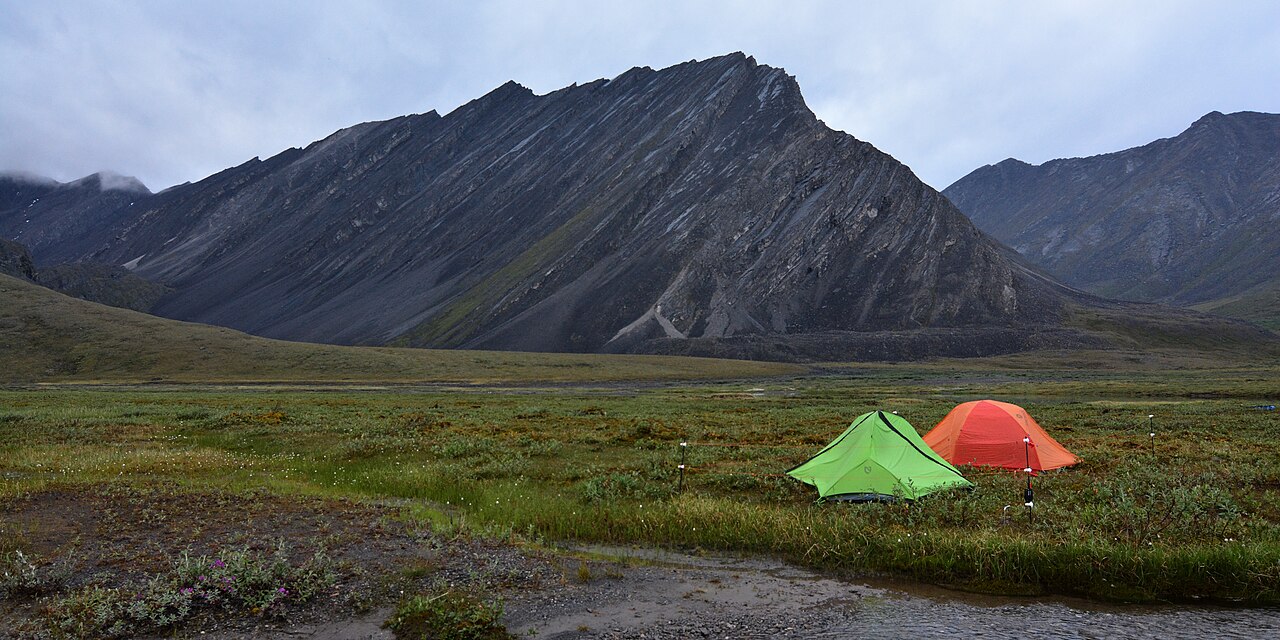 Two small backpacking tents pitched on open ground in a wide valley below a jagged rock ridge.