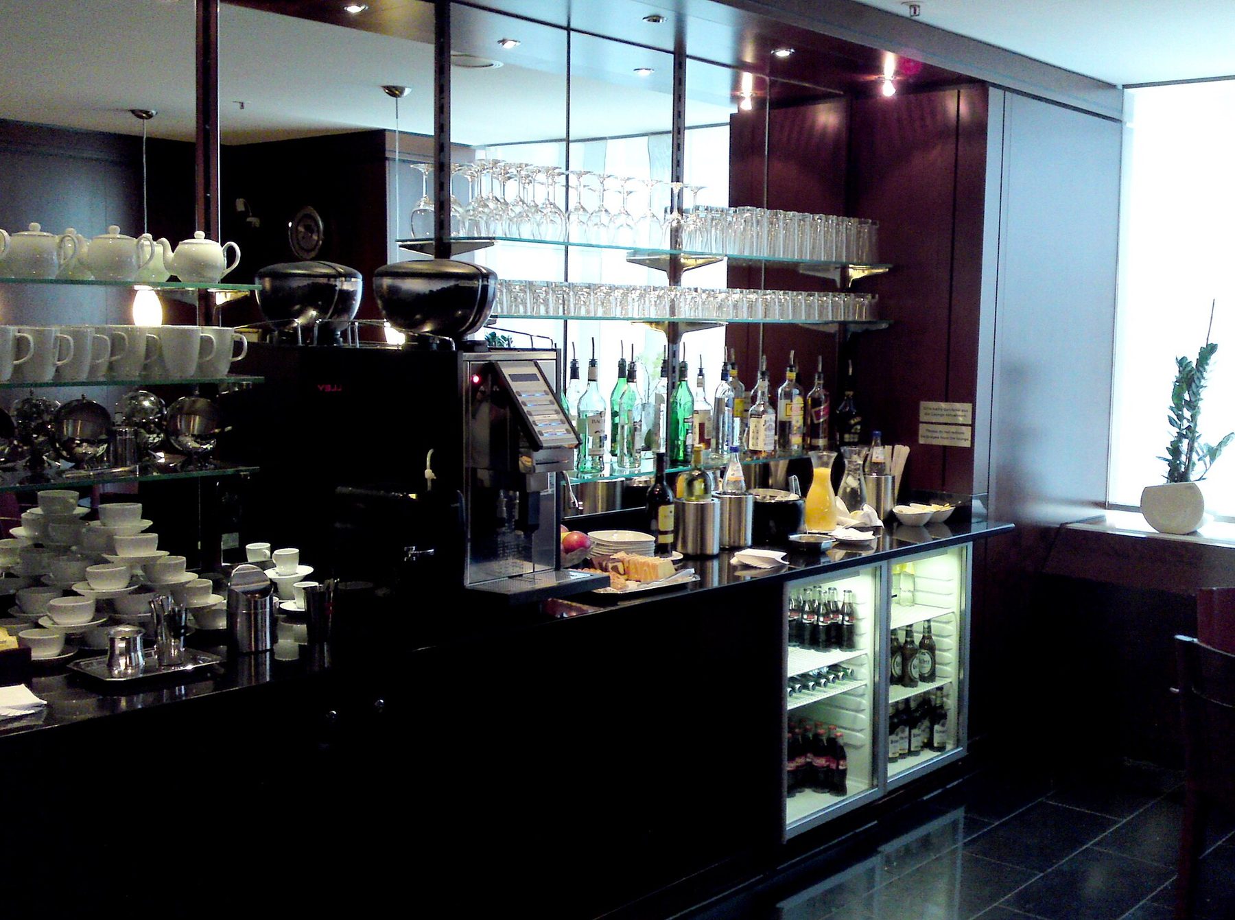 A quiet lounge with pale tiled flooring, low armchairs, a bar with high stools and an American Airlines Admirals Club sign