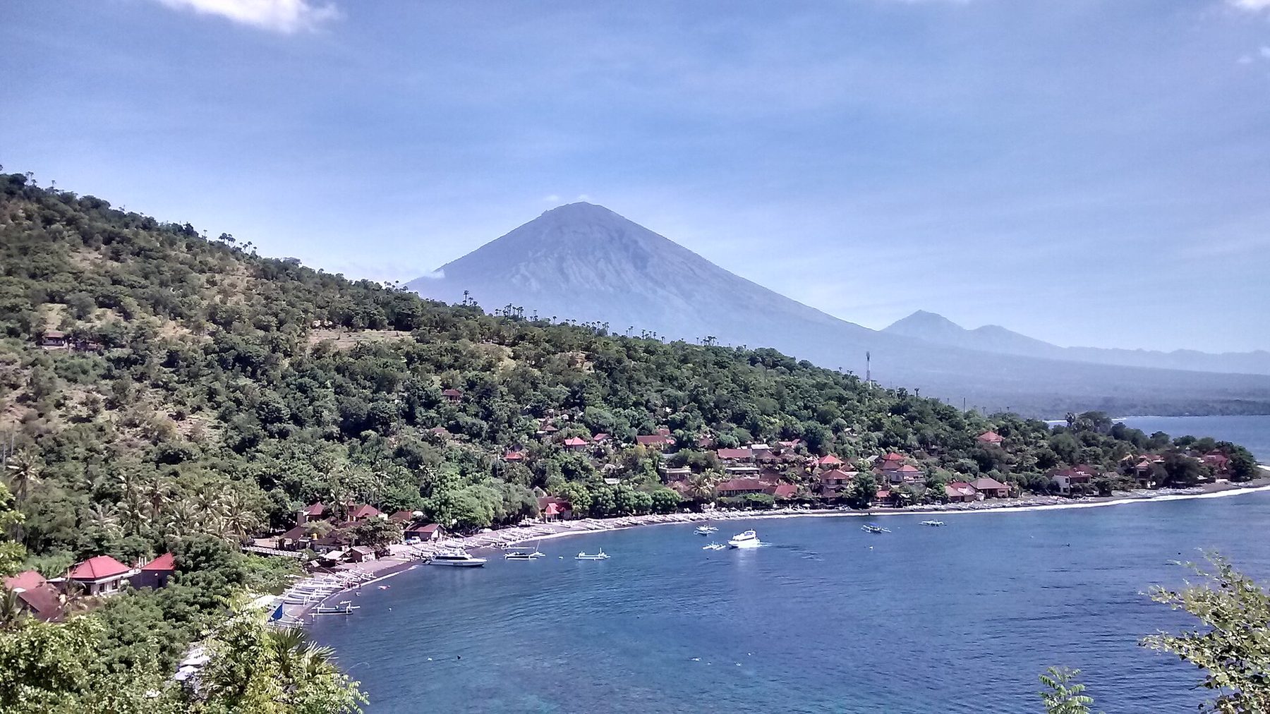 A dark volcanic shoreline at Bunutan in east Bali, with calm blue water and a green headland running out to sea