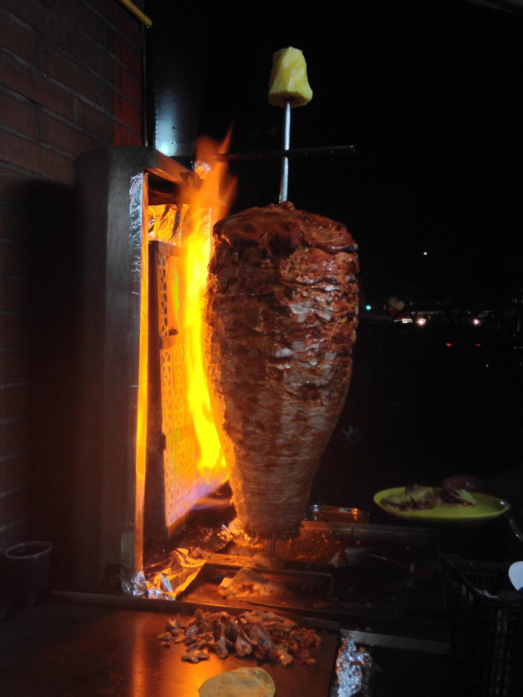 A cook slices meat from a tall cone of marinated pork on a vertical spit, with a pineapple on top and flames behind it