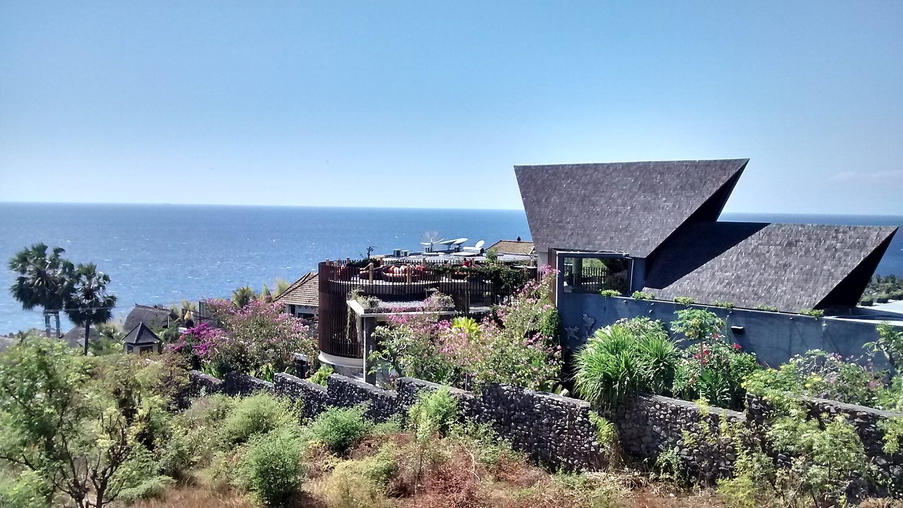 A hillside villa with steep angular shingled roofs above dry scrub, stone terrace walls and bougainvillea, with open sea beyond