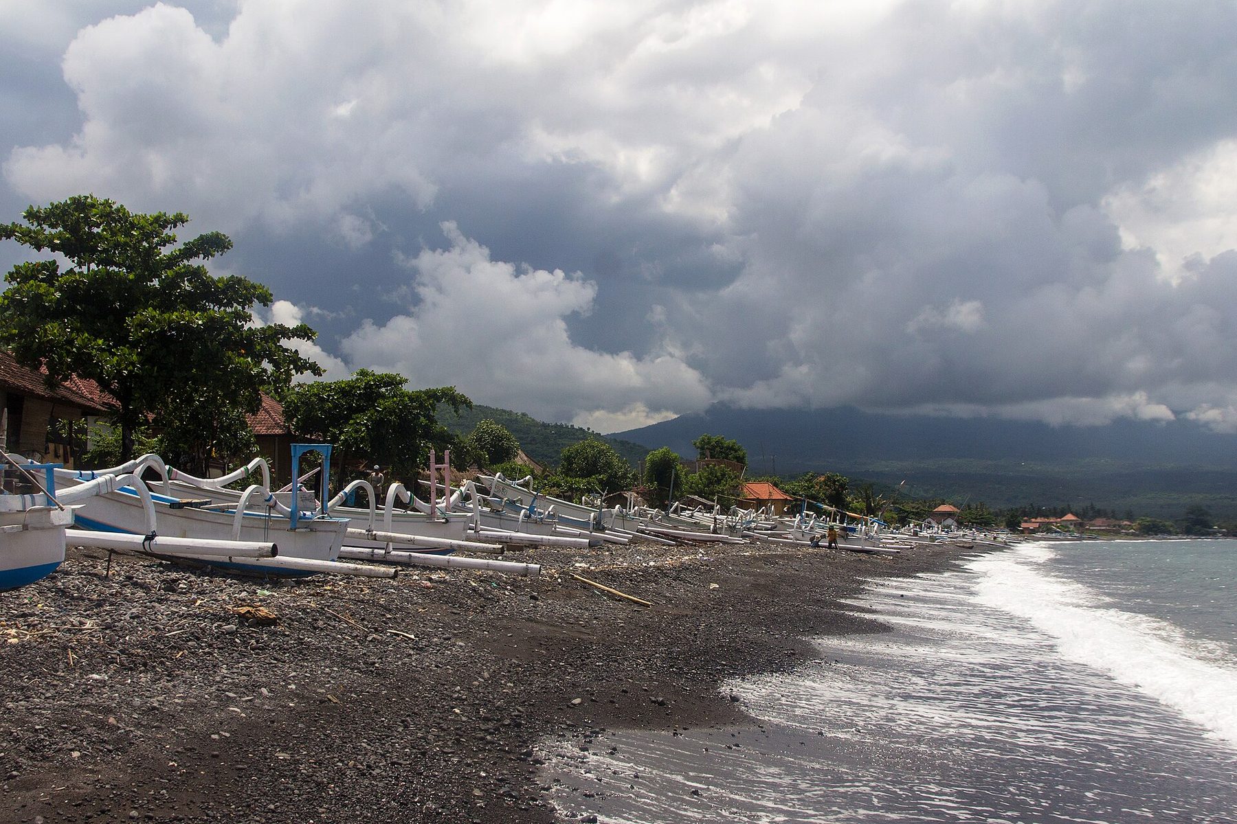 A long line of white and blue outrigger fishing boats on a dark pebble and black sand shore at Amed under heavy cloud, with a green ridge and a mountain flank behind the village