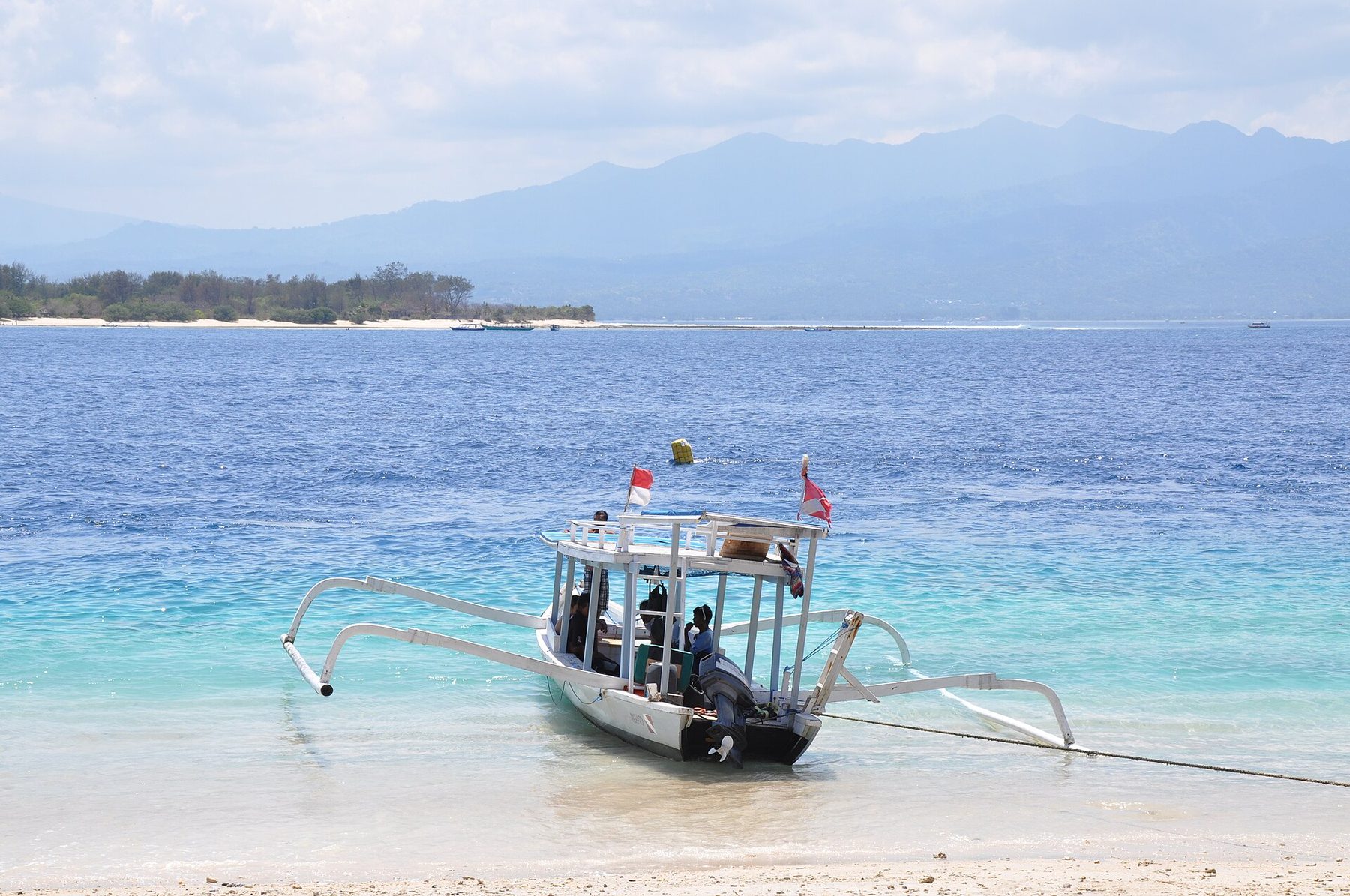 Clear turquoise shallows over pale sand off a low palm-lined shore with small moored boats
