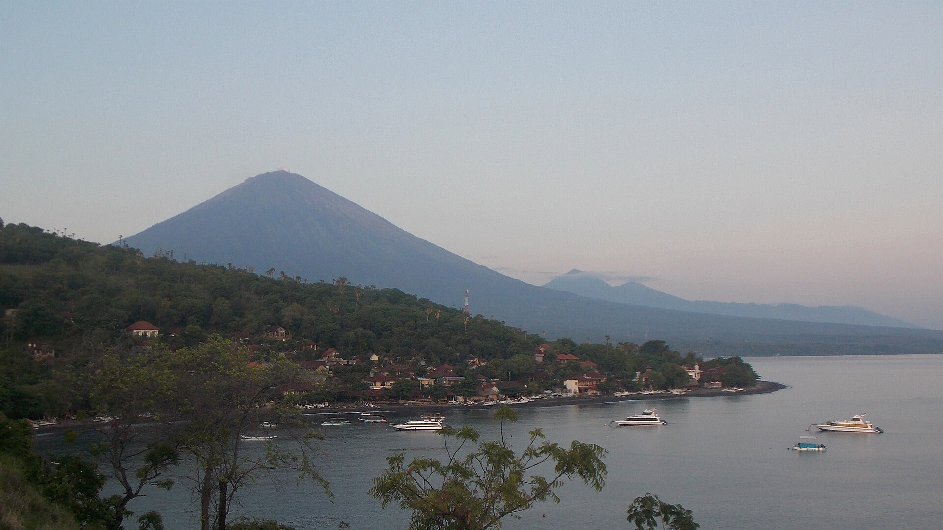 The cone of Mount Agung rising behind low green hills and village rooftops, seen from the coast at Amed
