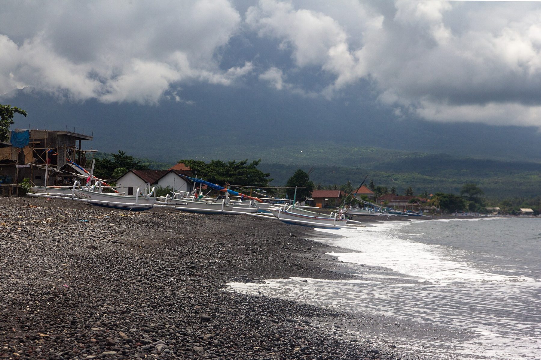 White outrigger fishing boats lined along a beach of black sand and grey pebbles under heavy cloud, with village roofs and a forested ridge behind