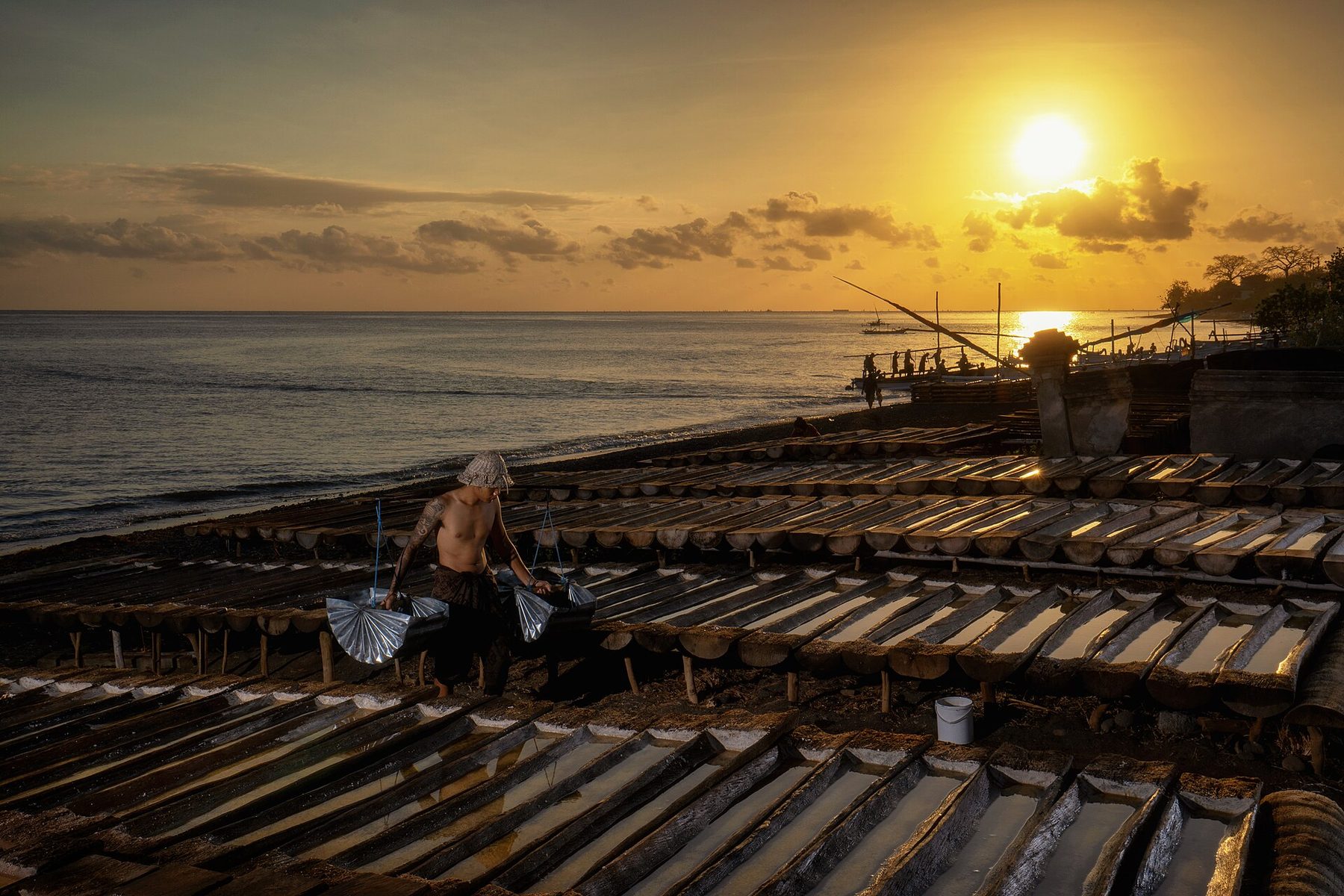 A man in a sun hat bends over a long wooden trough of seawater on a beach, working sea salt by hand