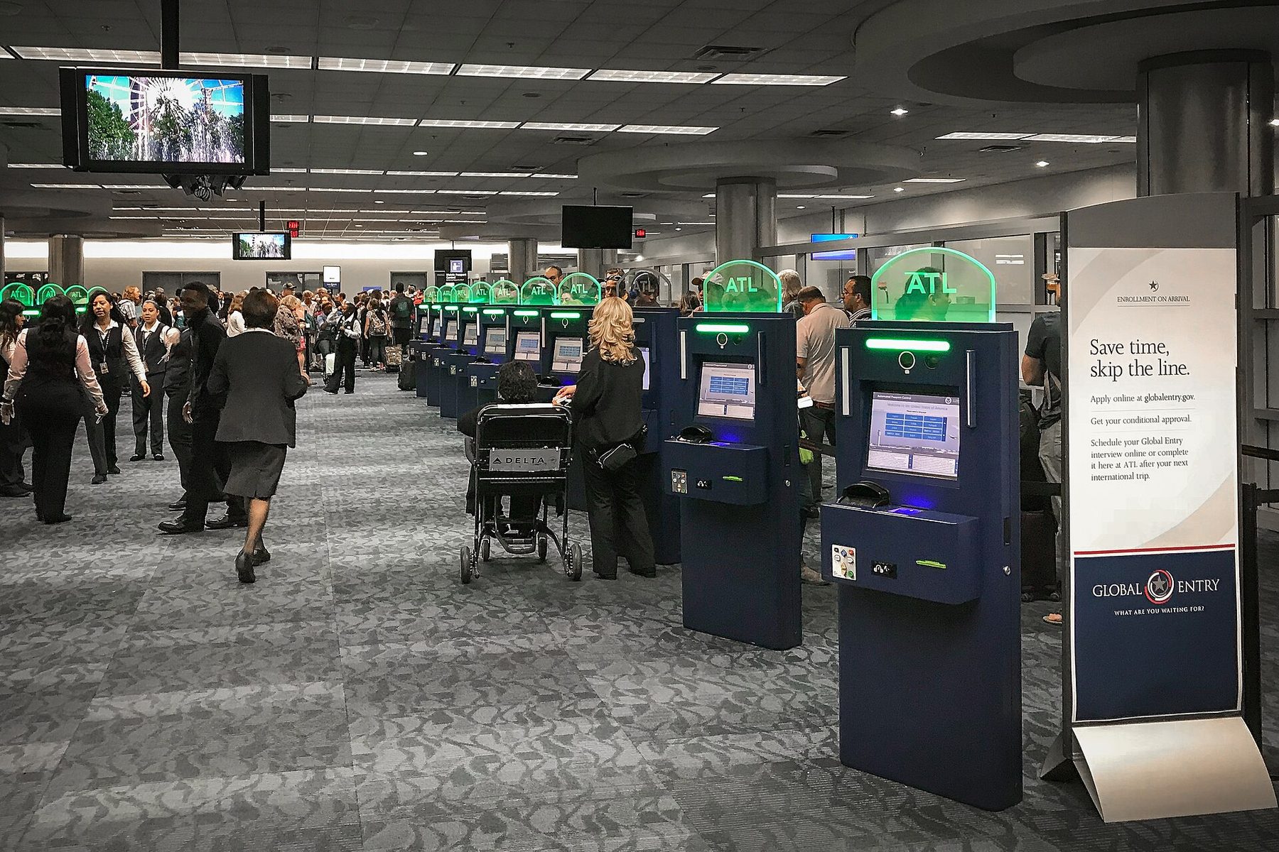 A row of Automated Passport Control kiosks with blue screens in an arrivals hall, travelers queuing behind a rope line