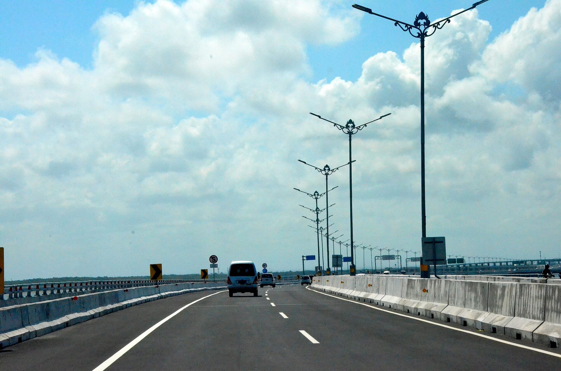A wide two-lane toll road with concrete barriers curves ahead under a blue sky, a white minivan and two cars in the distance, ornate street lamps lining the right-hand side