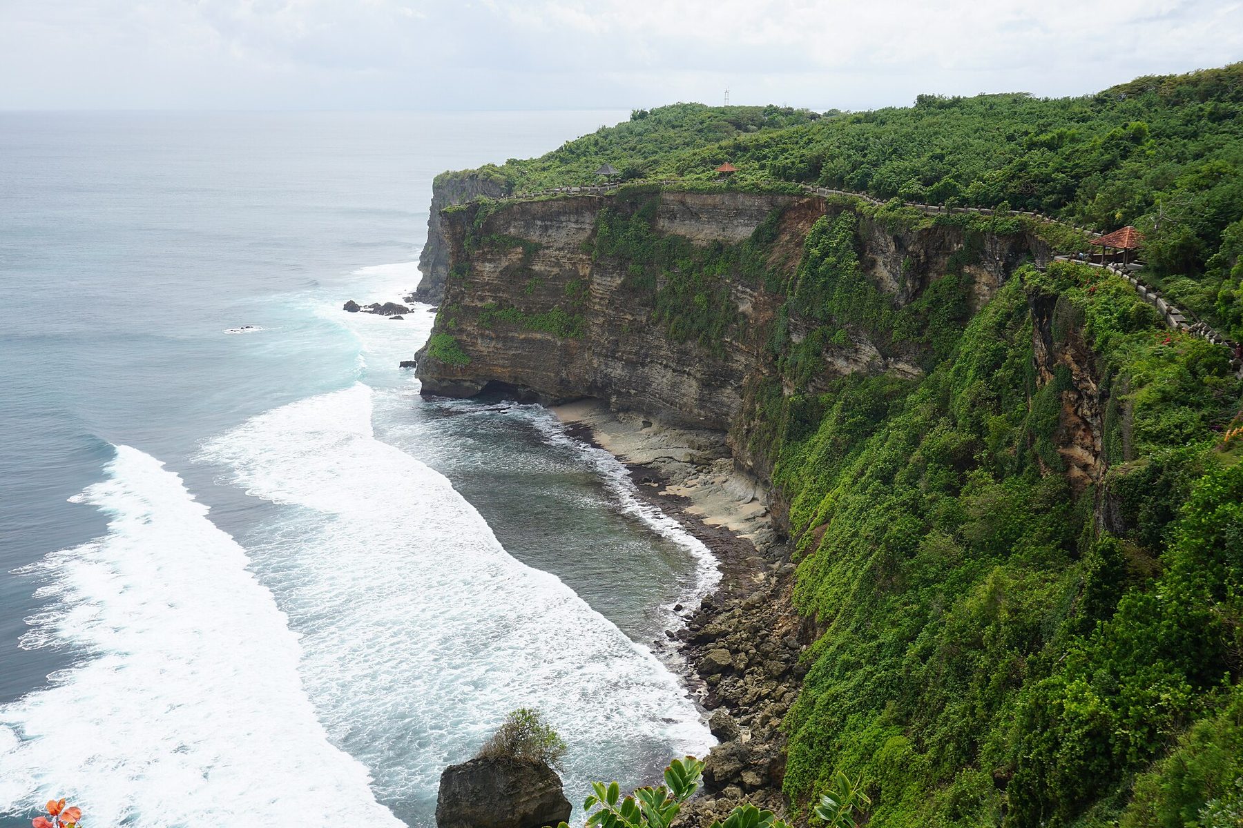 A green clifftop headland drops in tall limestone walls to a rocky shore where white surf lines roll in from an open grey-blue ocean, with a railed walkway and small pavilions along the cliff edge