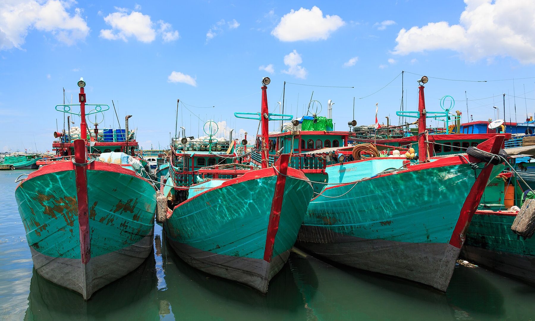 Three green and red wooden fishing boats moored bow-first in the water at Benoa Harbour under a bright blue sky