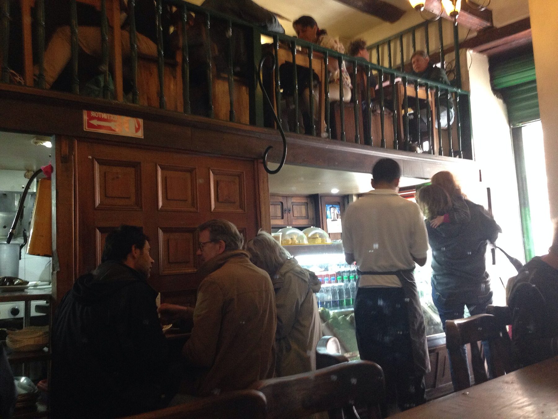 Diners at a dark wooden counter inside La Puerta Falsa, with a green wrought-iron balcony of tables above the ground floor, a lit display case and a drinks fridge behind the staff