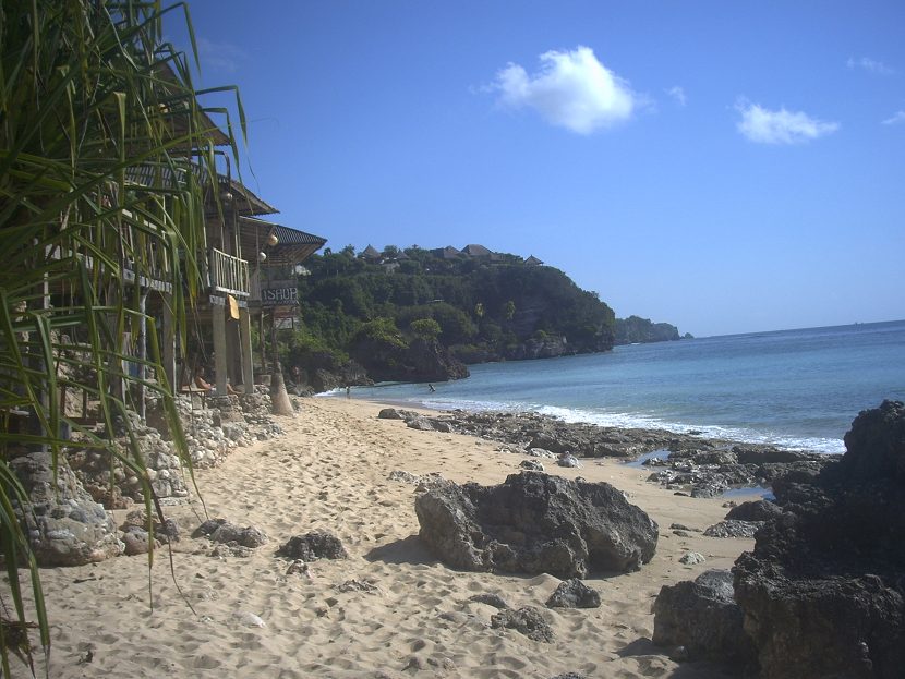 A small sandy cove below limestone cliffs with low buildings along the shoreline and pale green water breaking over a shallow reef
