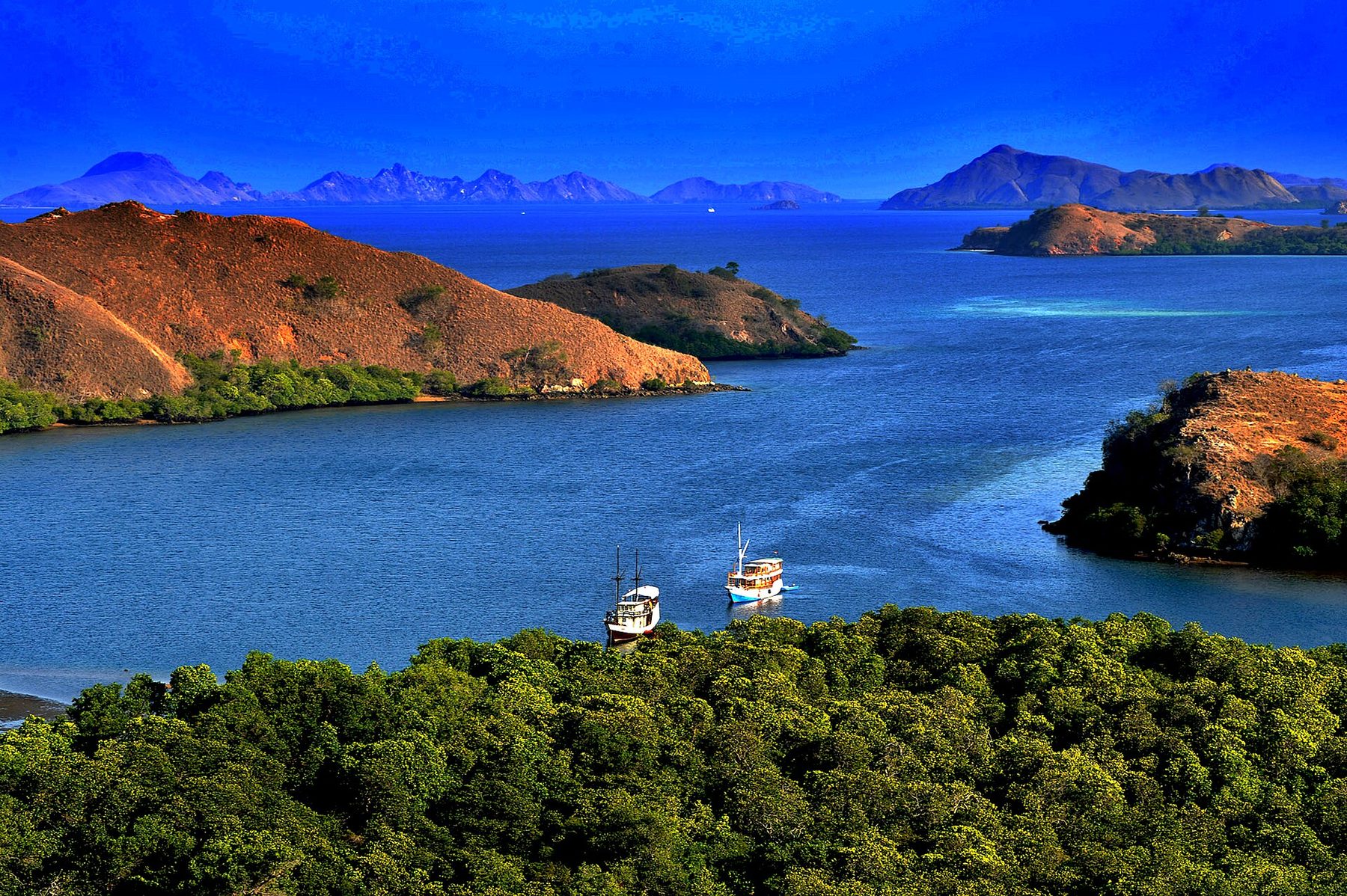 A wooden tourist boat at anchor in a calm bay off Rinca Island, with dry brown hills rising behind it