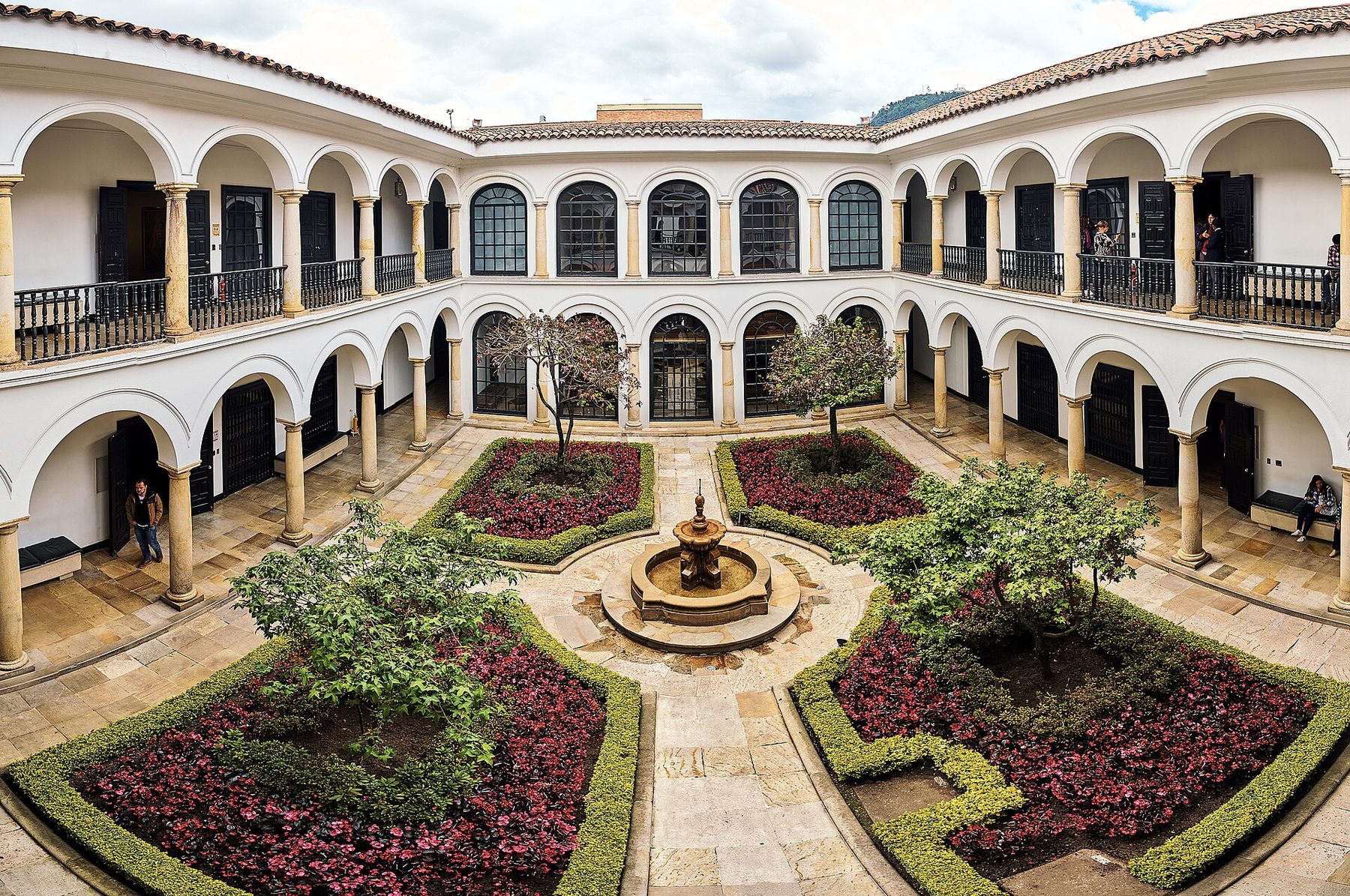A two-storey white colonial courtyard with arcades and a tiled roof, a stone fountain at the center and four beds of red and green planting around it