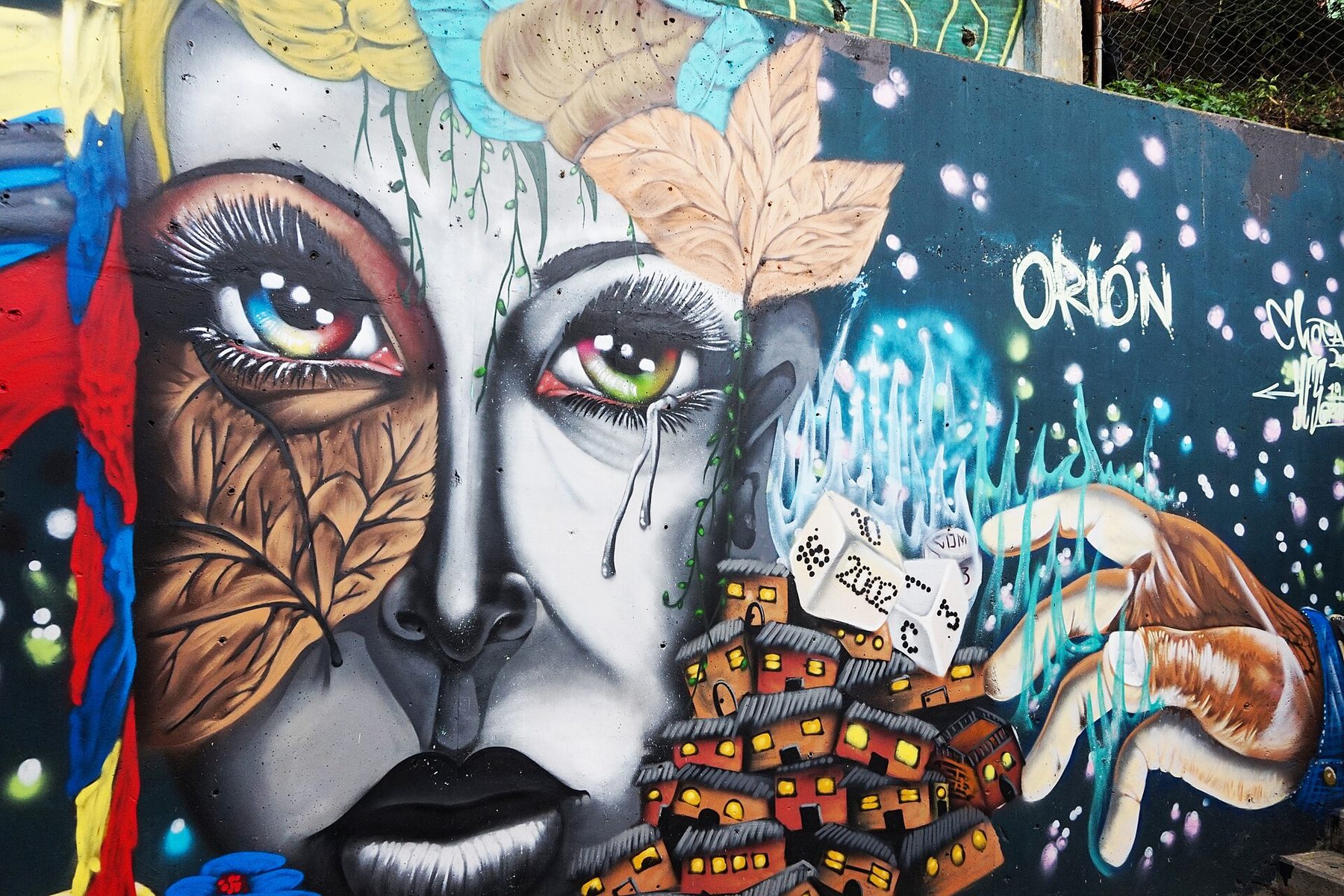 A painted wall mural of a large face with leaves across one cheek, a hillside of small orange houses and dice marked 2002, with the word Orión lettered beside it