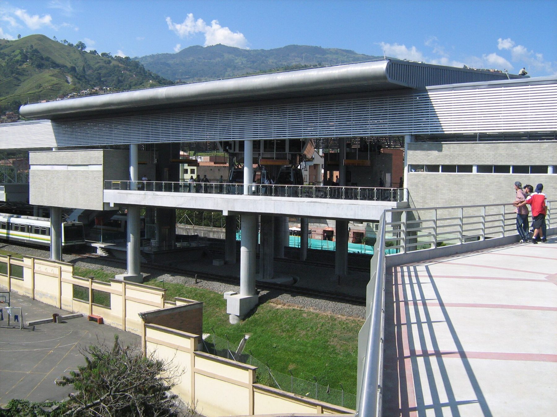San Javier Metro station in Medellín, with a train on the lower platform, cable cars docked on the upper level and green hills behind