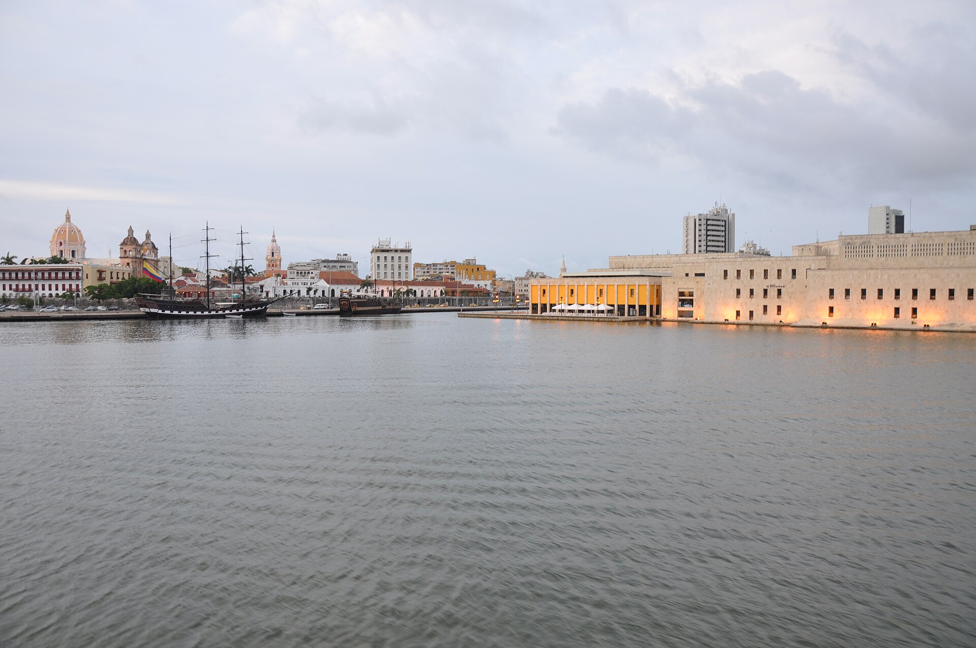 The Cartagena waterfront at dusk across the bay: church domes and low colonial rooftops on the left, tall sailing-ship masts at the quay, and the floodlit stone convention center on the right