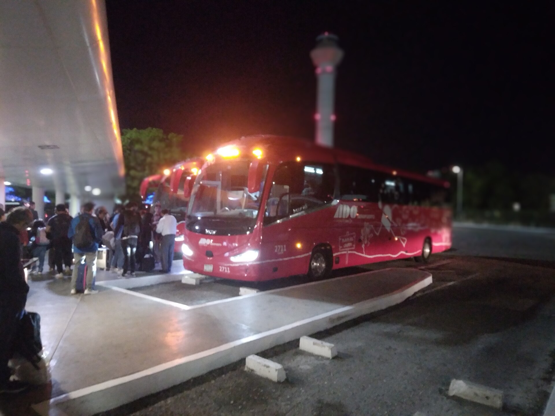 A red ADO coach stands at a covered boarding platform at night, headlights on, while a line of passengers with suitcases waits on the kerb and the lit control tower rises behind
