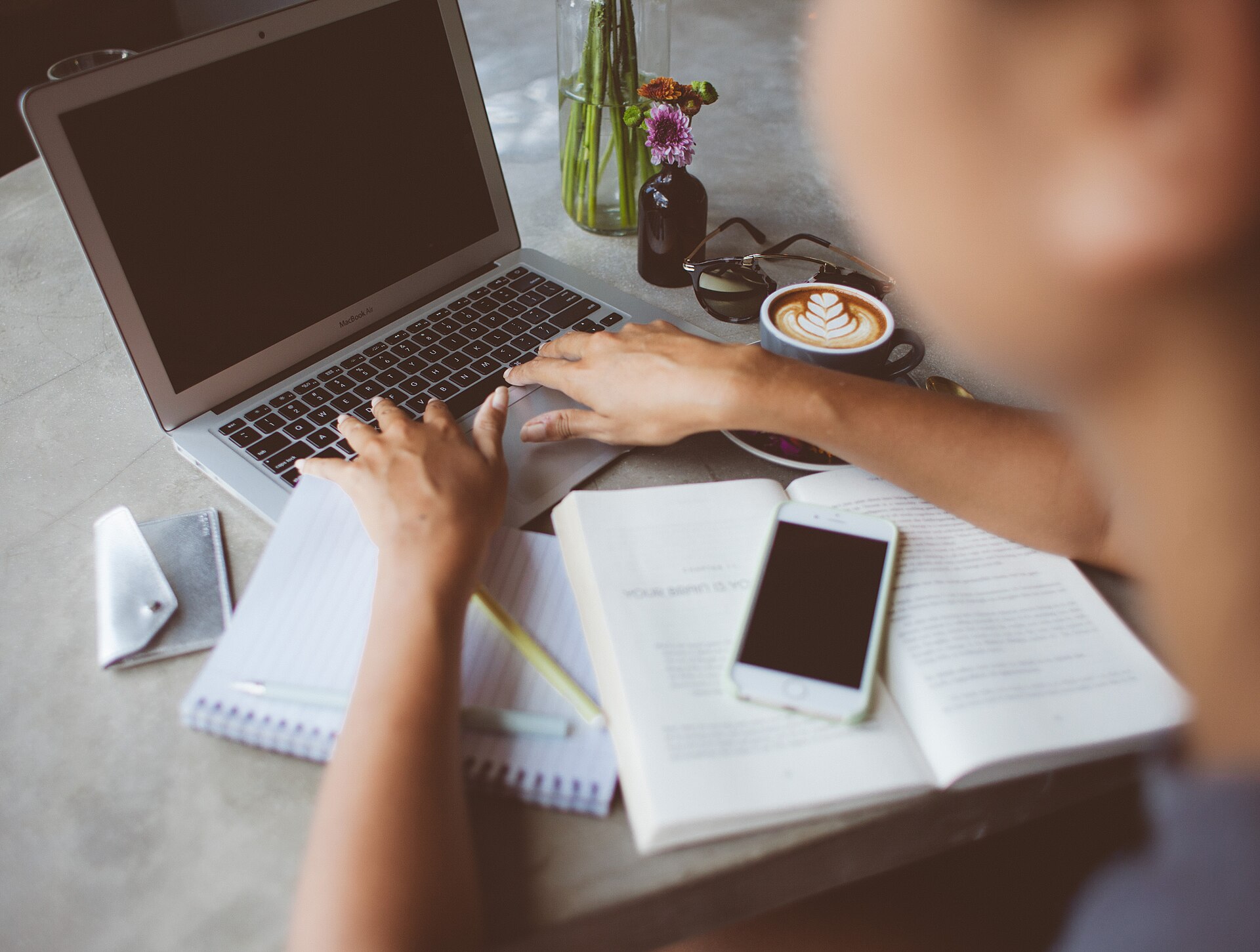 Seen over a person's shoulder, two hands type on a laptop at a concrete cafe table beside a latte, an open book, a spiral notebook, a phone and sunglasses
