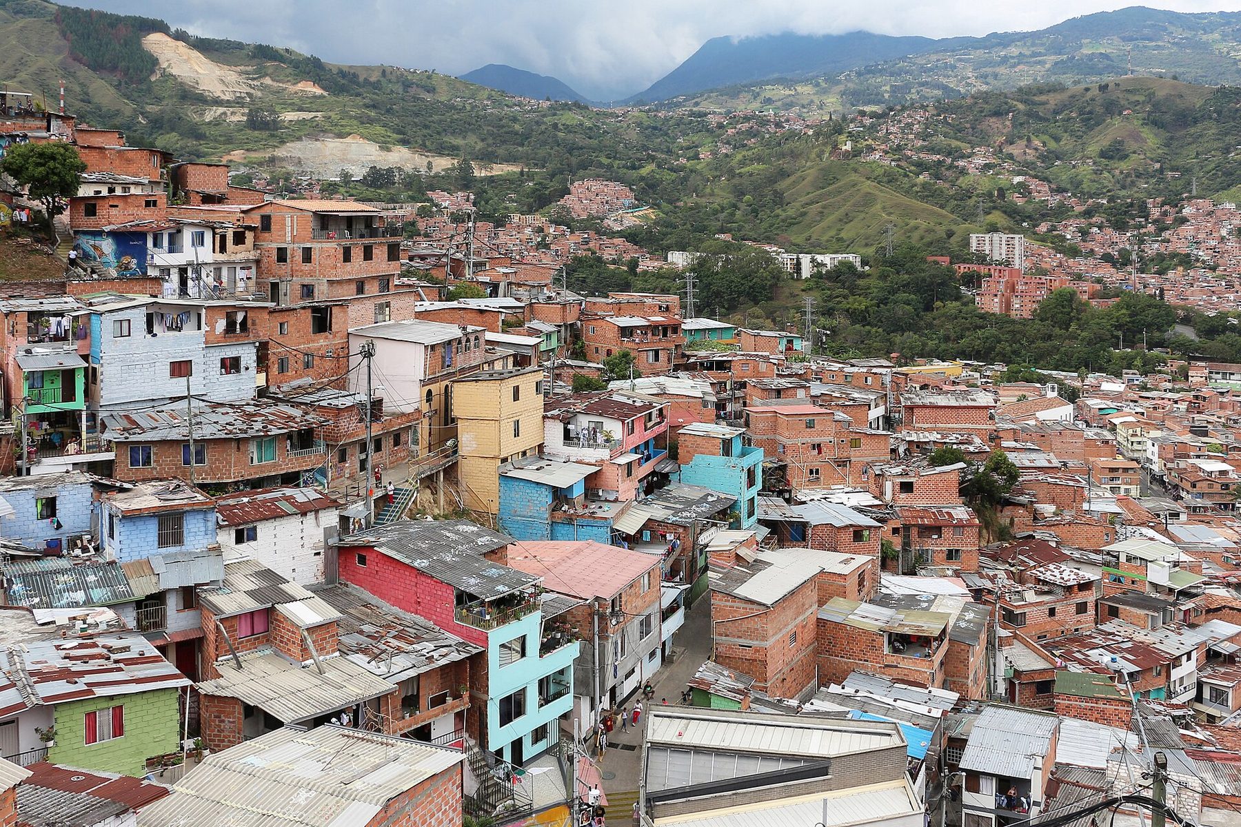 Brick and painted houses stacked up a steep hillside in Comuna 13, Medellín, with green mountains and the wider Aburrá valley behind them