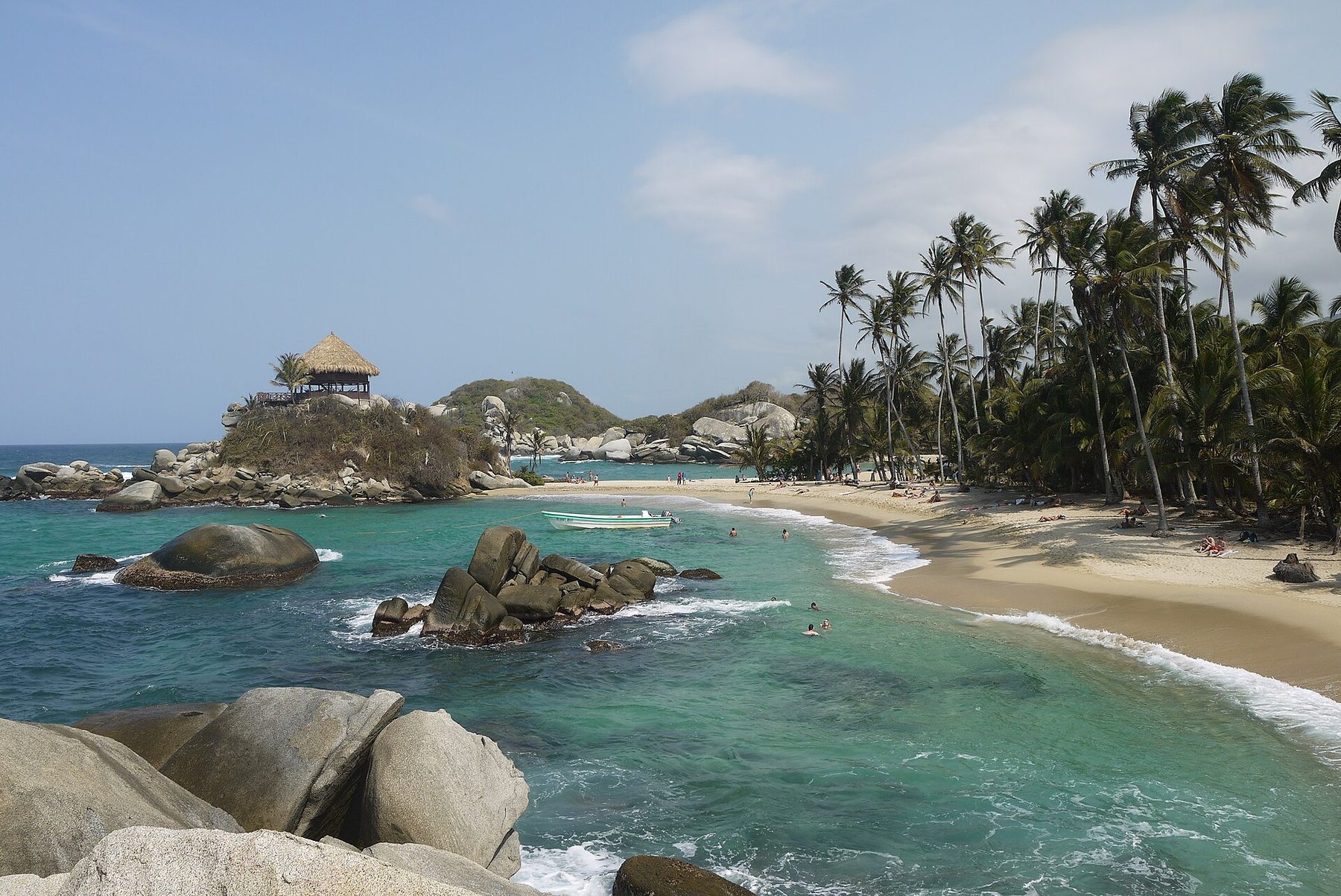 A curved sand beach at Cabo San Juan del Guía in Tayrona National Natural Park, with turquoise water, granite boulders, tall palms and a thatched lookout hut on a rocky headland