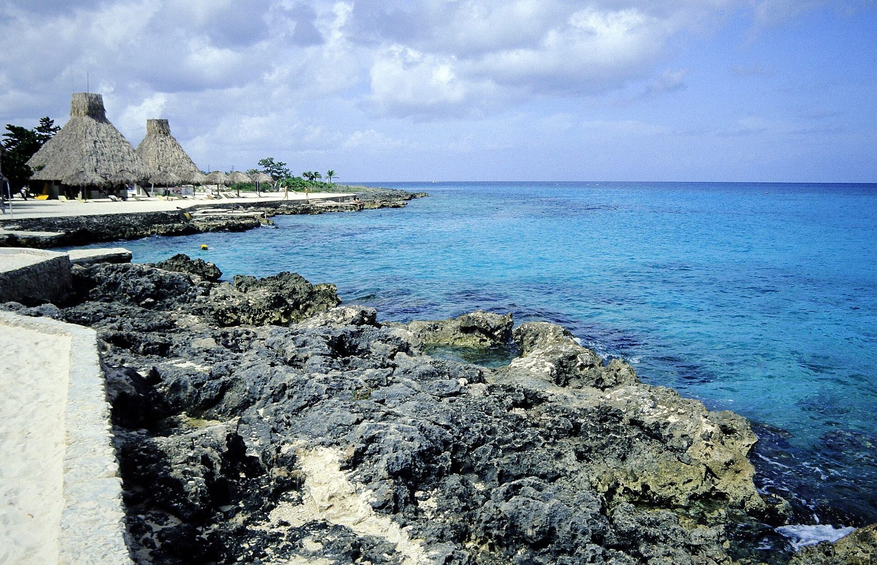 A rocky shoreline at Chankanaab park with a wooden pier, thatched palapas and pale turquoise water in an inlet
