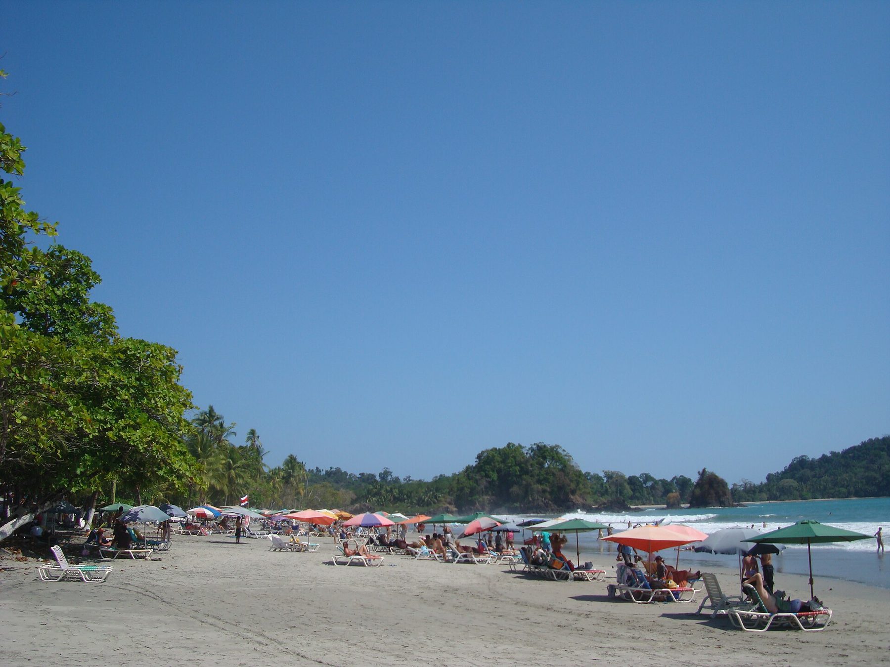 A curved sand beach inside a forested cove, with calm shallow water and green headlands on both sides