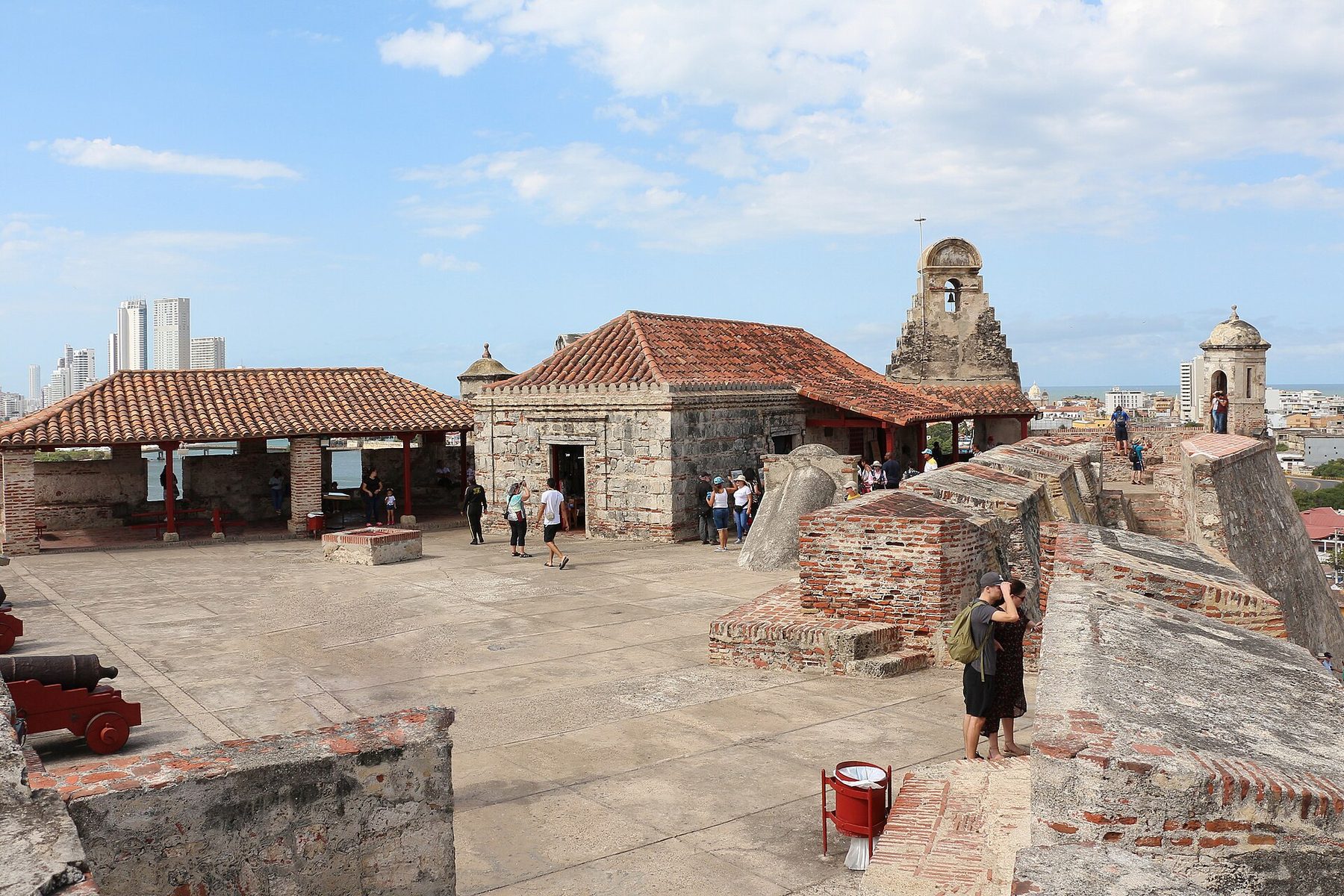 The upper terrace of Castillo San Felipe de Barajas in full sun, with a cannon, tile-roofed shelters, a stone bell tower and visitors, and Cartagena beyond.