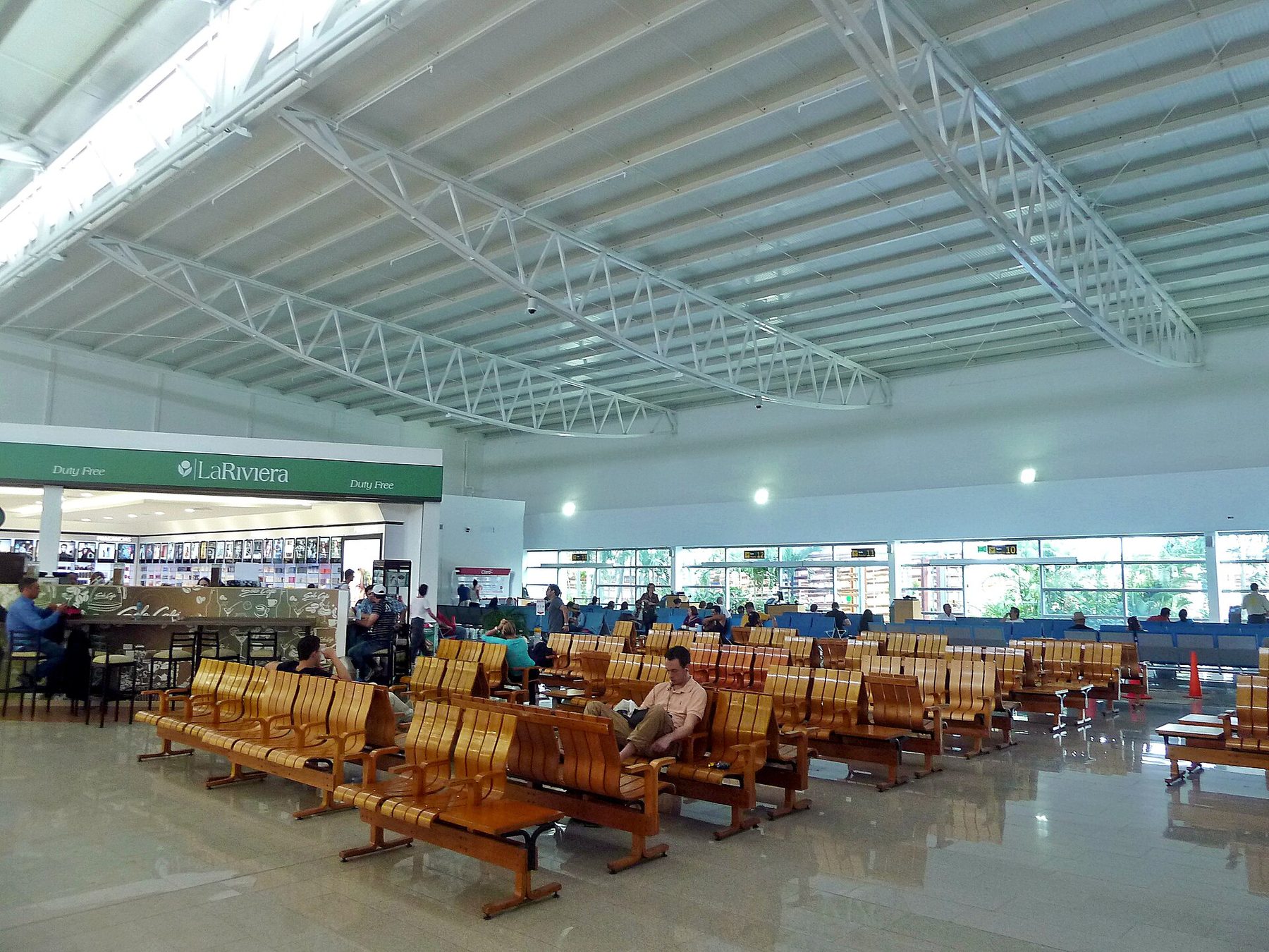Passengers waiting in rows of wooden bench seats in the bright departure lounge at Rafael Núñez International Airport in Cartagena, with a duty-free shop along one wall