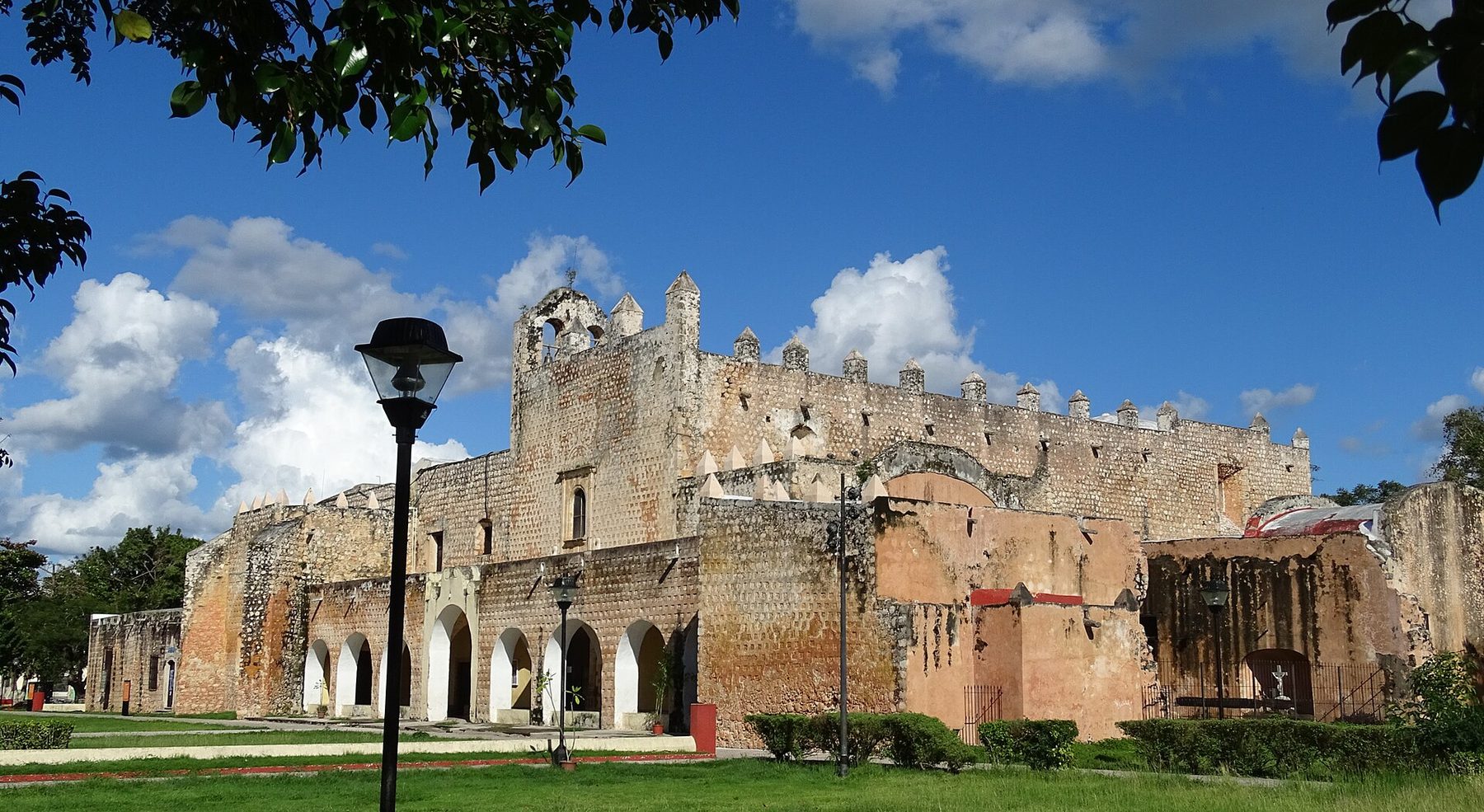 A long pale stone church and monastery with a bell gable and arcaded walkway stands behind a low wall under a clear sky