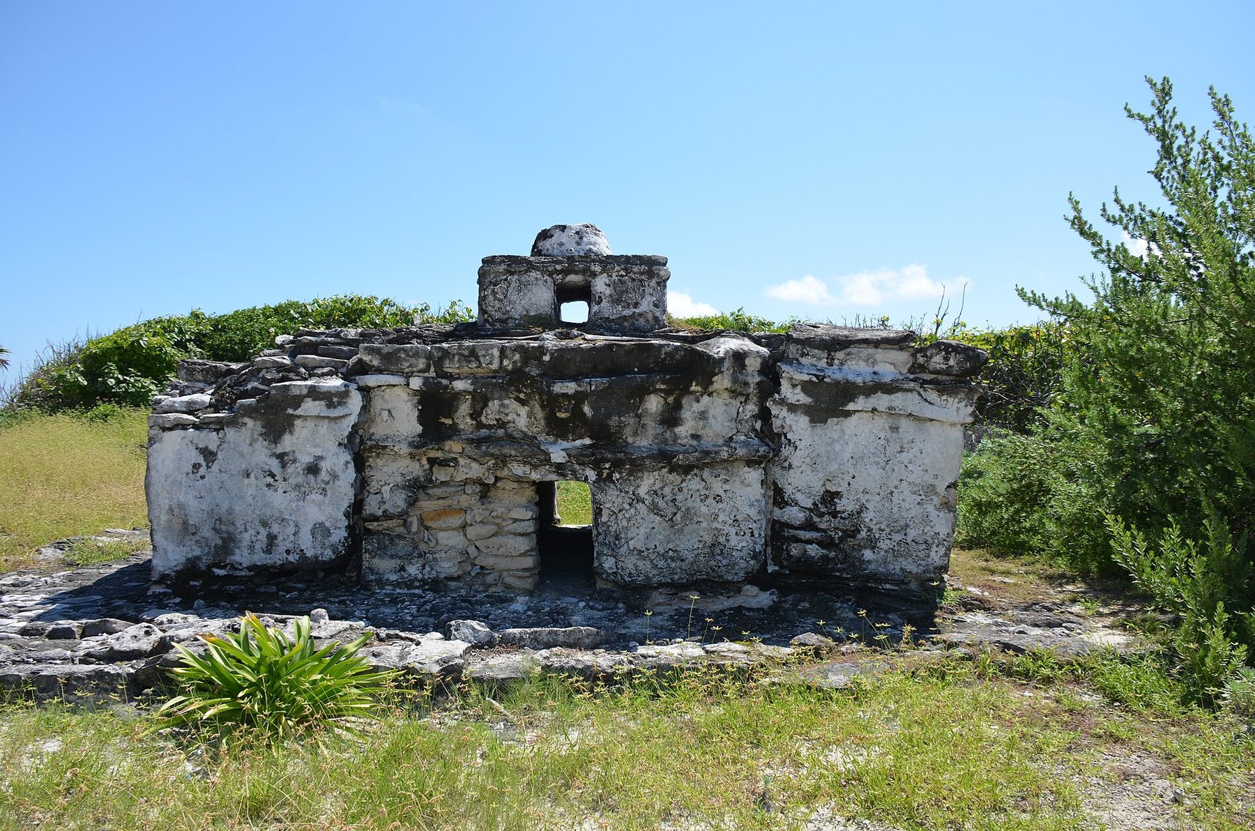 A low rectangular stone ruin with a small rounded turret on its roof, standing on grass and scrub under a bright blue sky