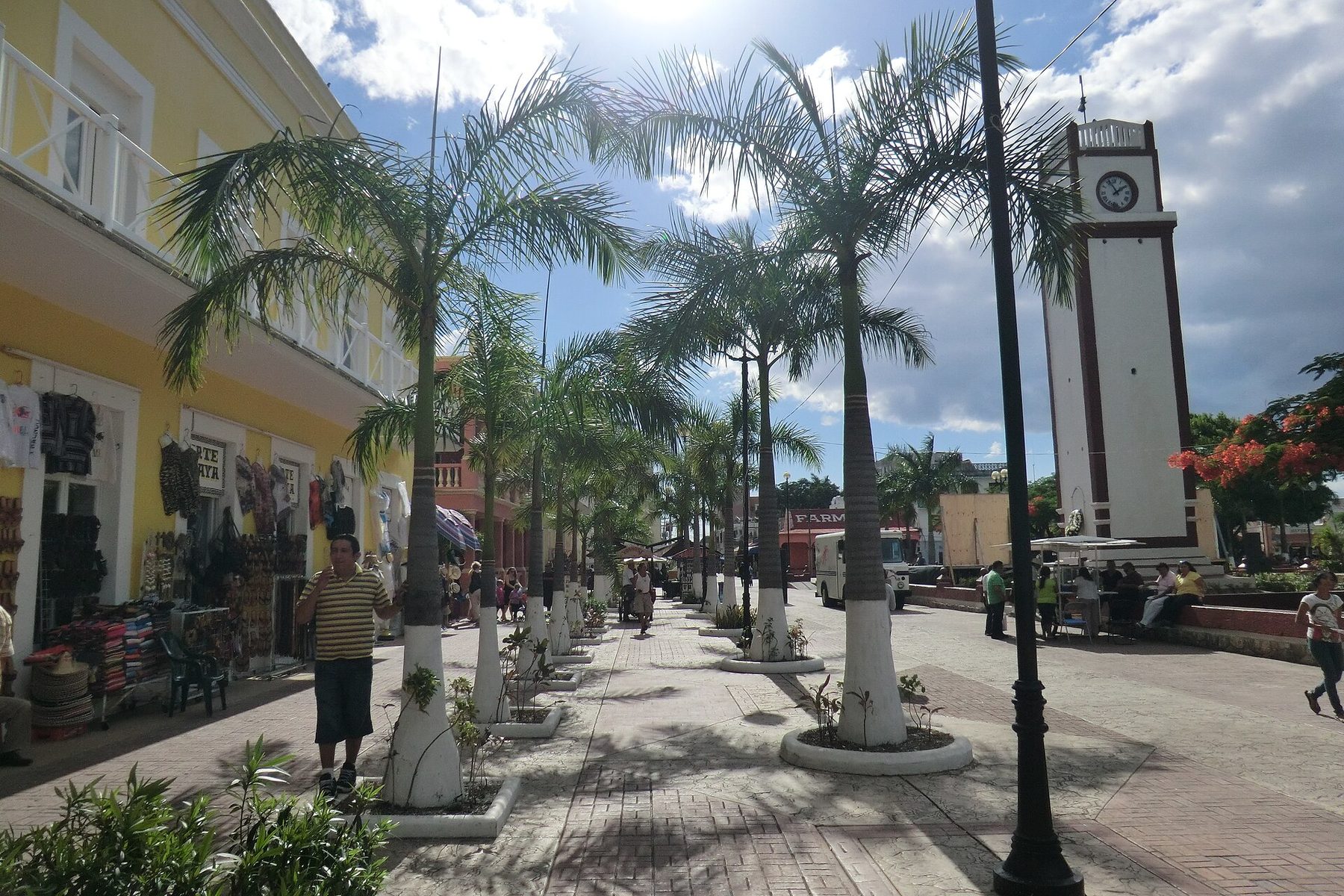 A palm-lined pedestrian street in central San Miguel de Cozumel with shops selling hats and clothing on the left and a white clock tower on the right