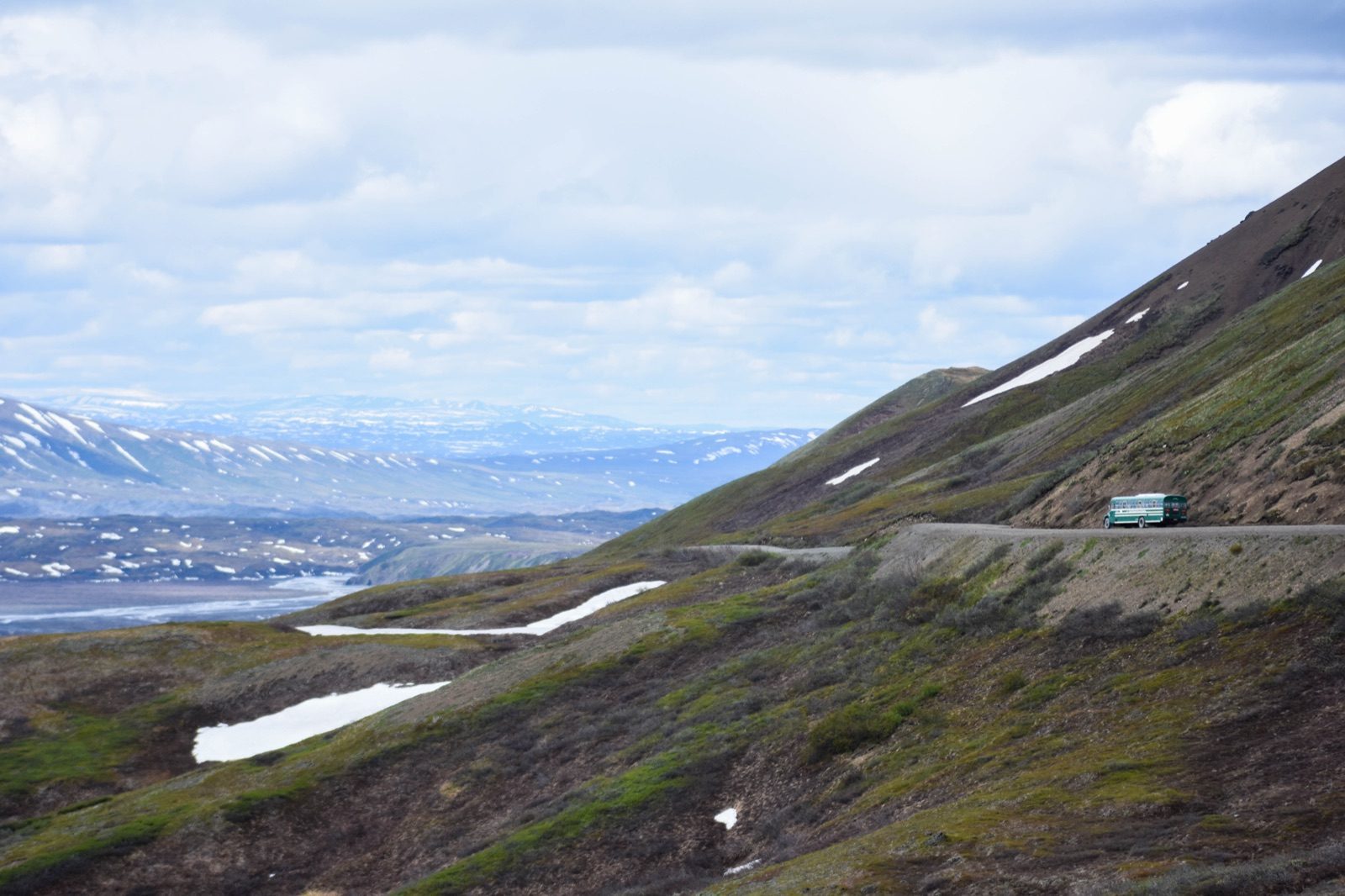 A green park bus drives along the unpaved Denali Park Road as it winds along a mountainside