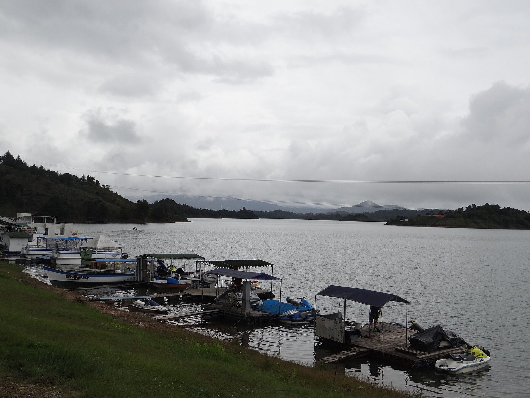 A waterfront walkway in Guatapé lined with moored tour boats, with wooded hills across the reservoir behind them