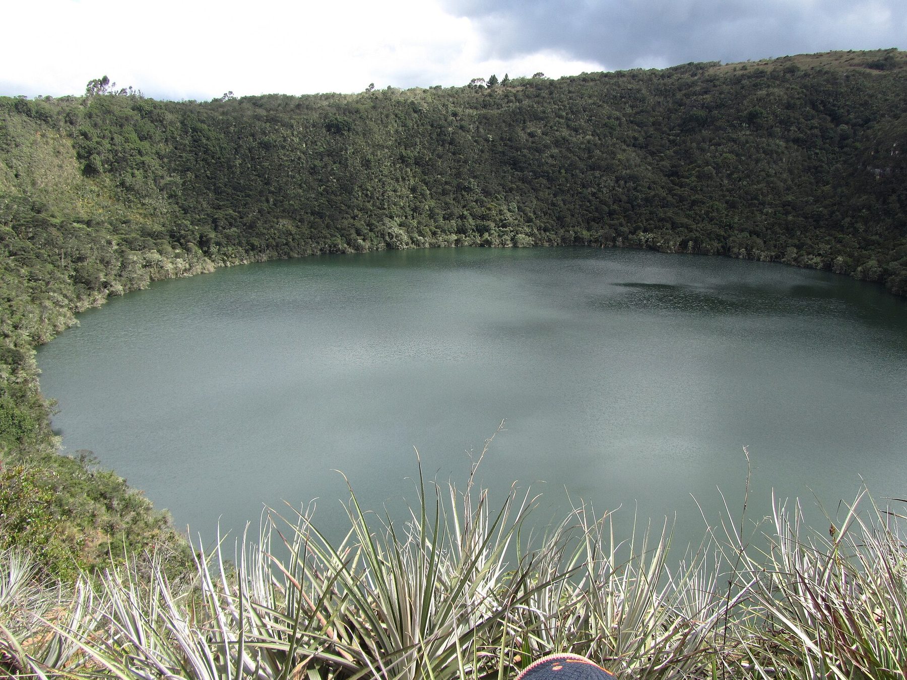 A near-circular lagoon of dark green water held in a bowl of bare and forested hills, seen from a rise above the shore