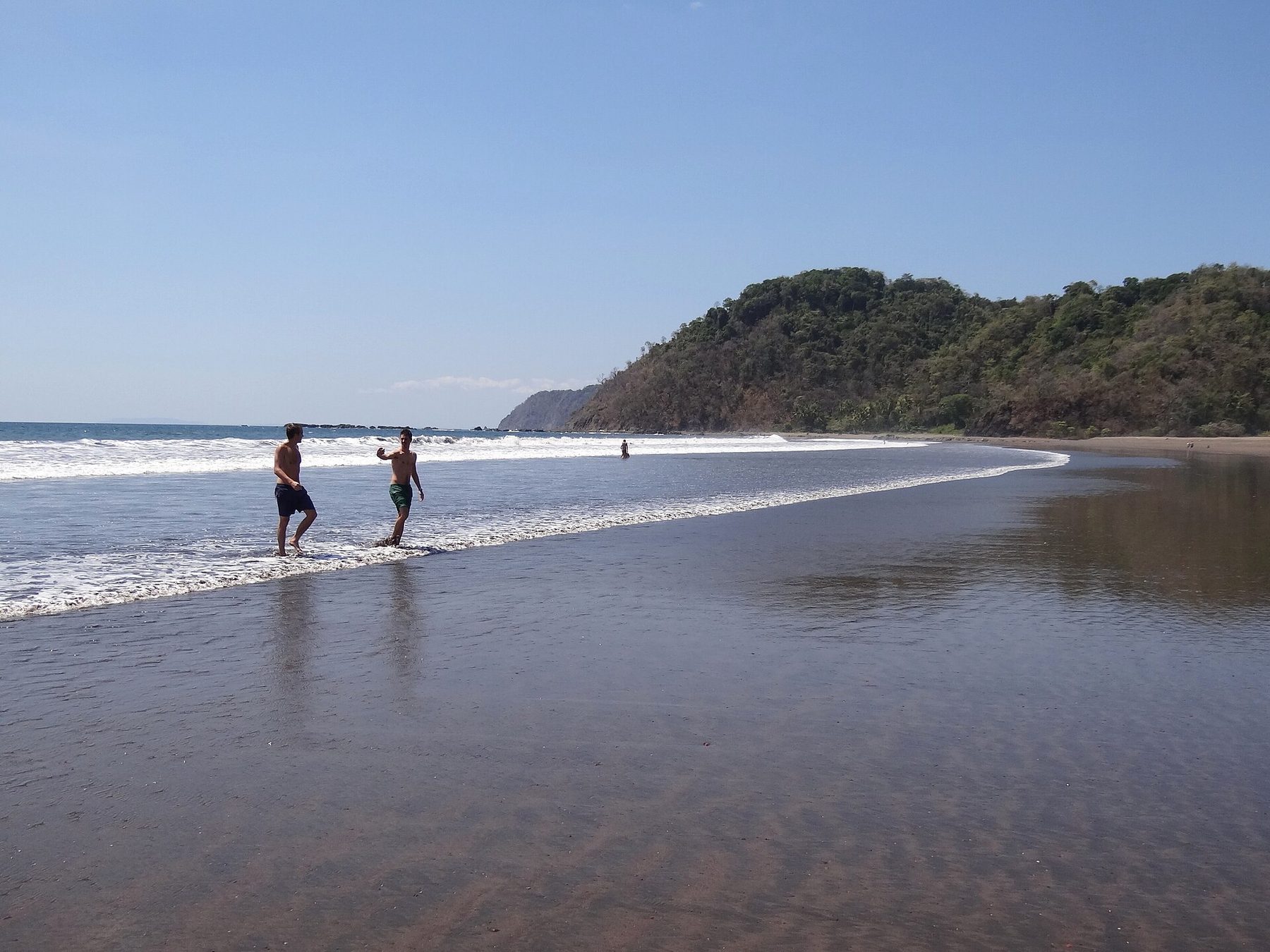 Swimmers and surfers stand in shallow water along a wide Pacific beach under a bright sky, with green hills at the far end