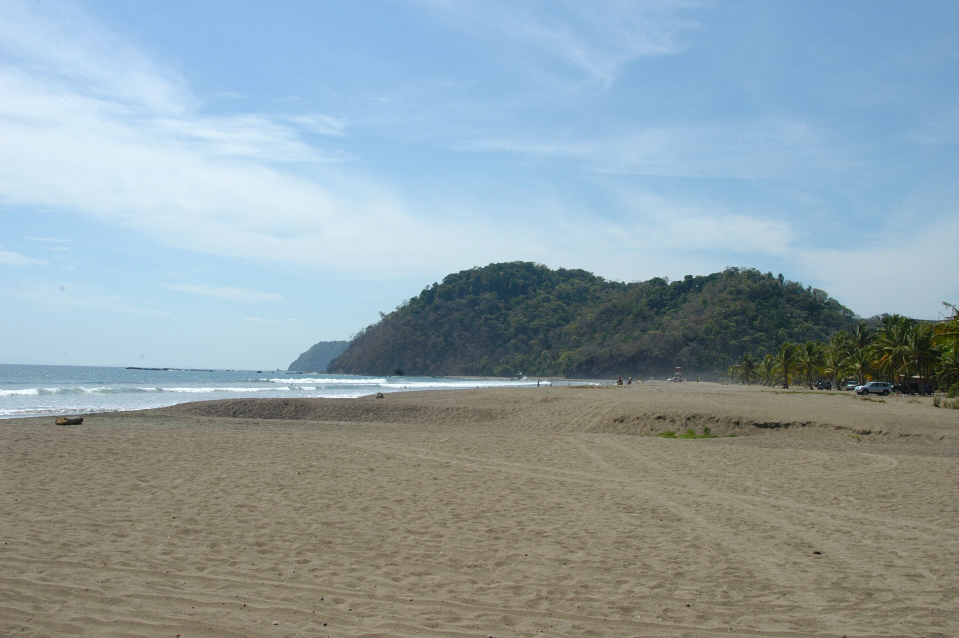 A broad stretch of pale sand at Jaco Beach with small waves breaking in lines offshore, palms and parked cars along the treeline, and a green headland at the end of the bay
