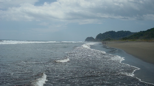 Grey surf running up the dark sand at Playa Hermosa under a cloudy sky, with a few figures far down the beach and a wooded ridge behind