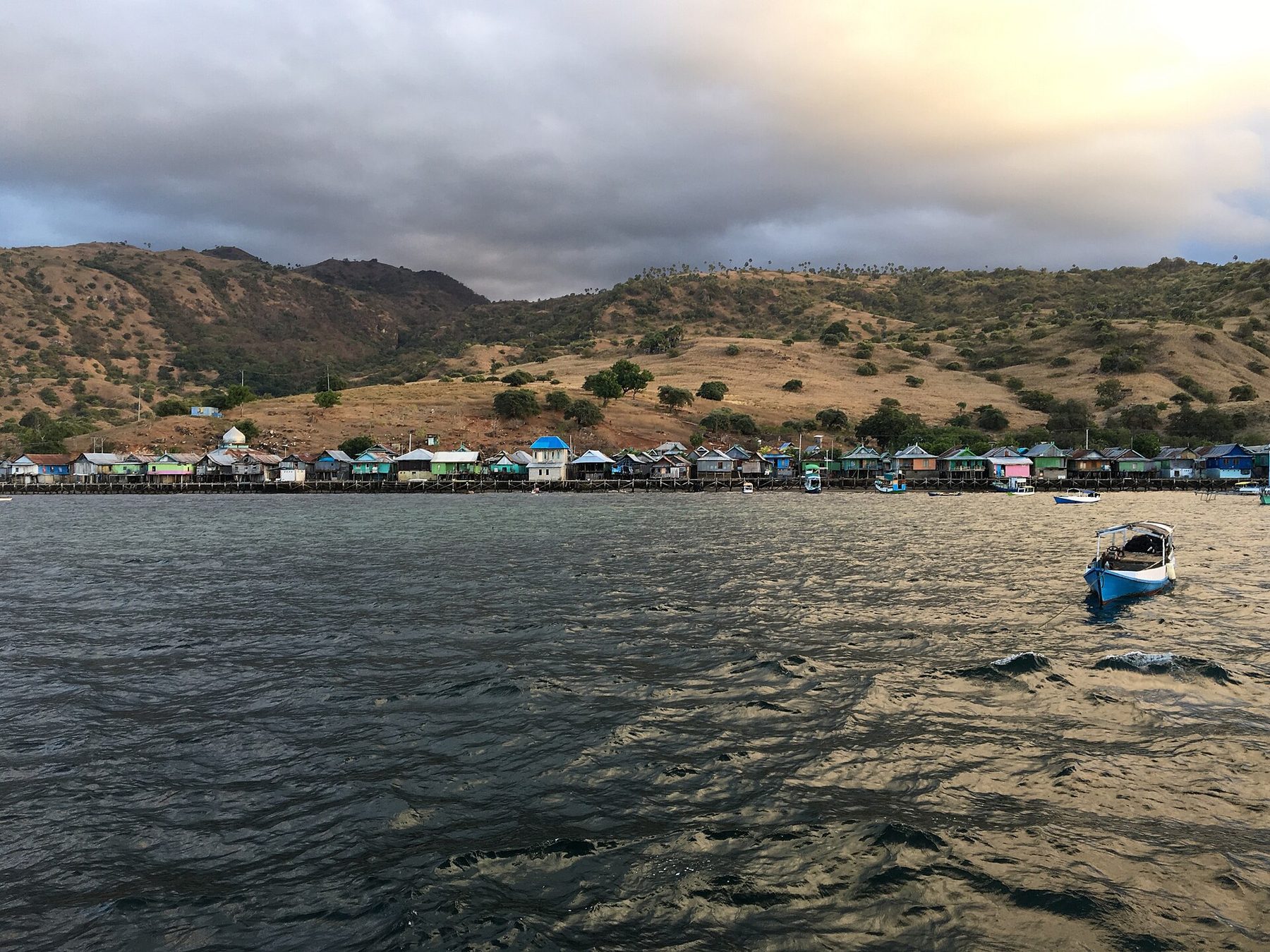 Wooden and concrete houses on stilts along a shoreline below a bare brown ridge, with small boats moored in the shallows