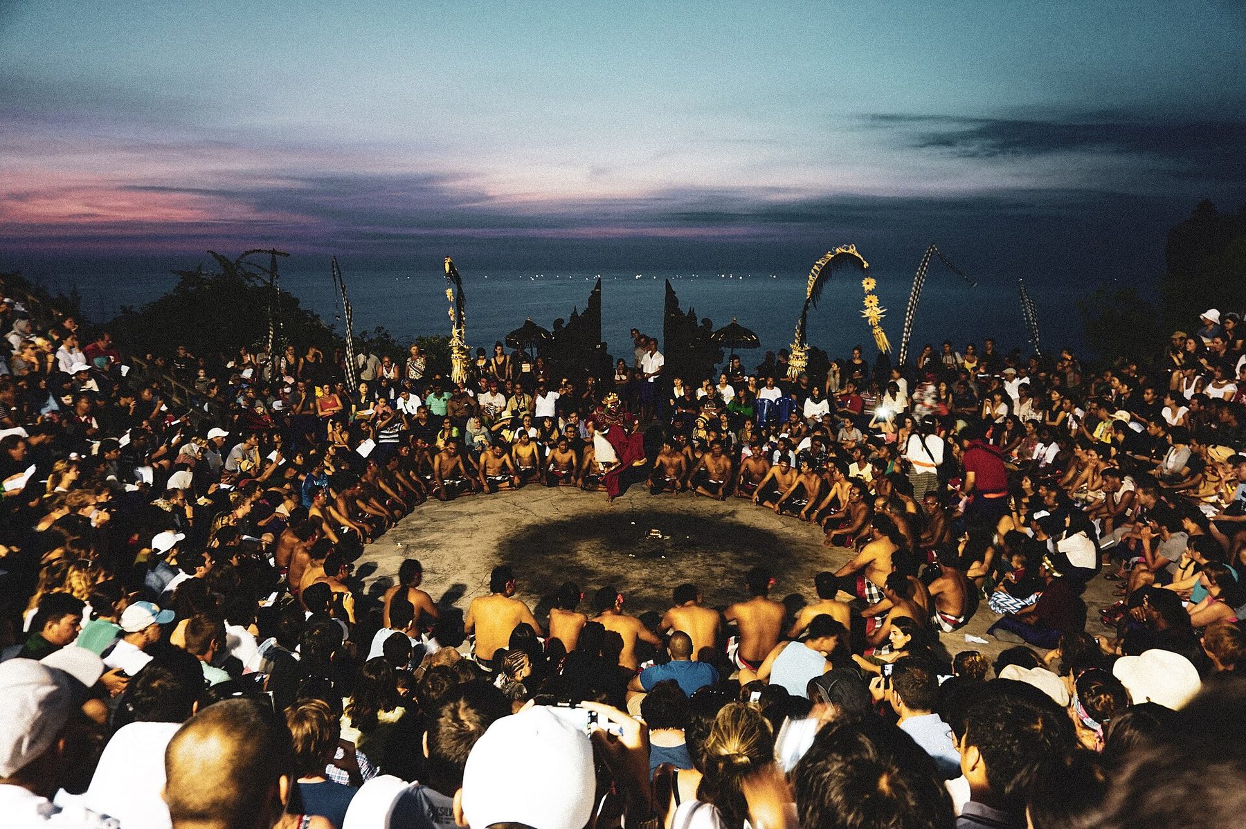 A ring of seated chanters with bare torsos and checked cloths around a burning fire on the open-air stage at Uluwatu, the sky darkening behind them