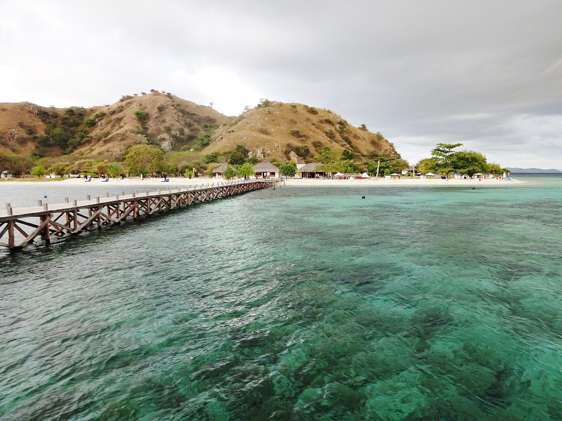 A long wooden jetty runs over clear green shallows to a white beach on Kanawa Island, with thatched buildings under trees and dry brown hills behind
