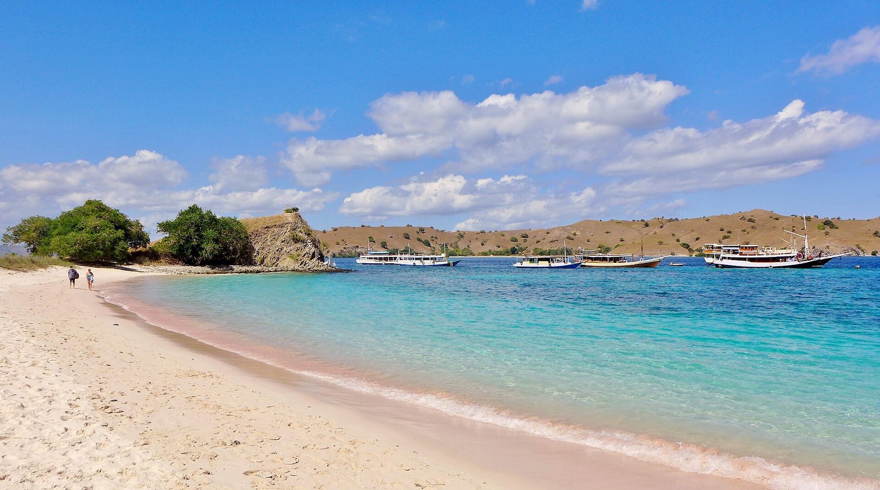 A curve of pale sand with a pink tinge at the waterline meets clear turquoise shallows, with wooden tour boats anchored offshore and dry brown hills beyond