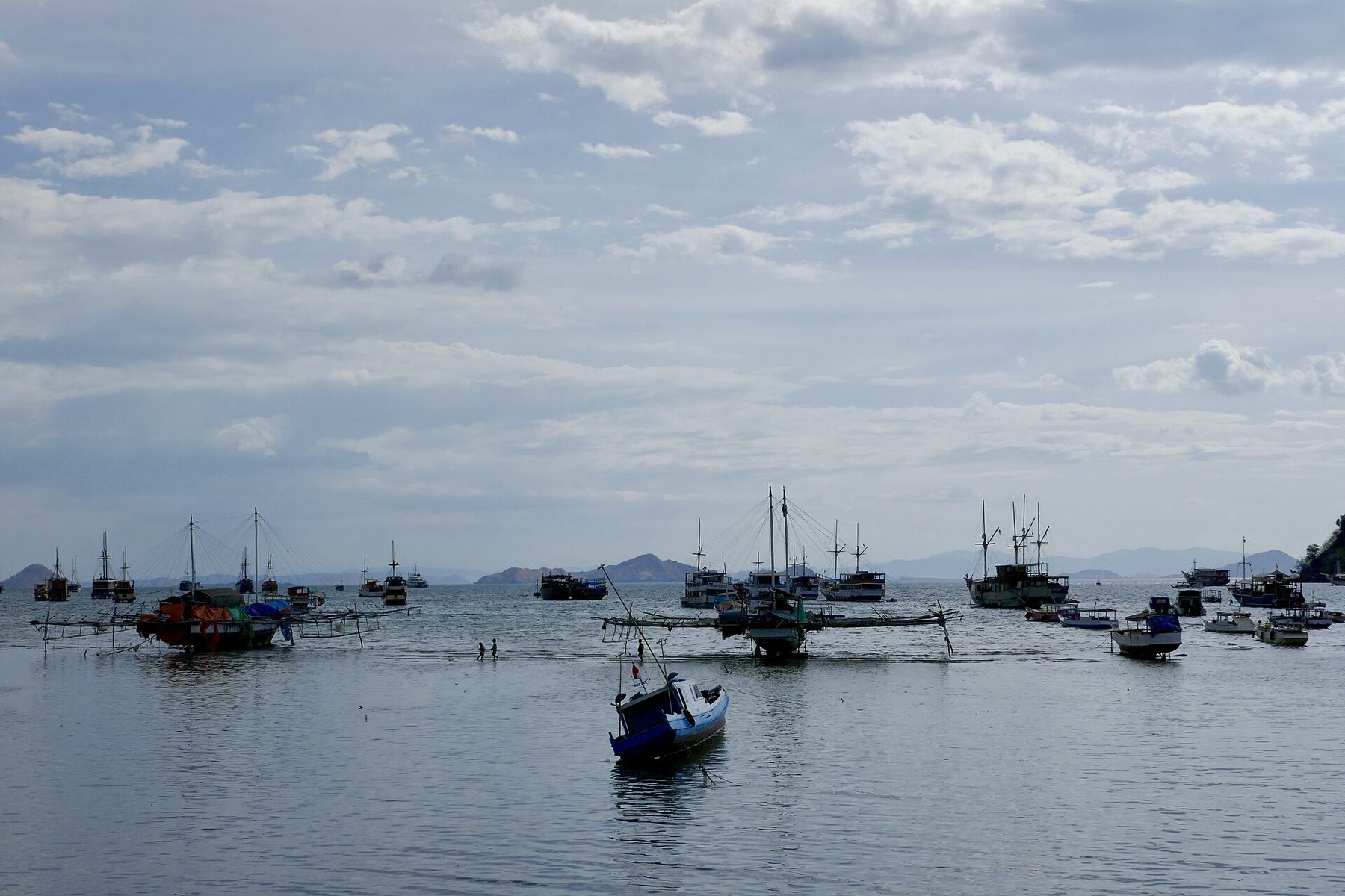 Dozens of wooden boats lie at anchor in a calm bay in front of low green hills