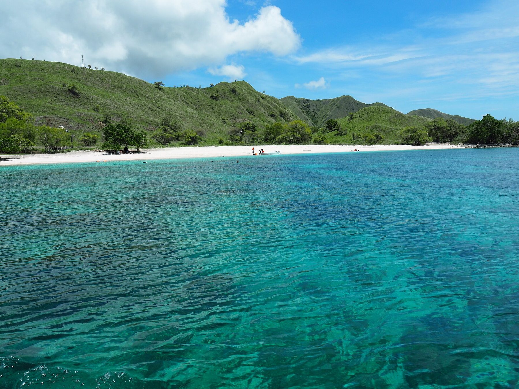 A curving pale beach meeting clear turquoise shallows, with green hills rising behind it.