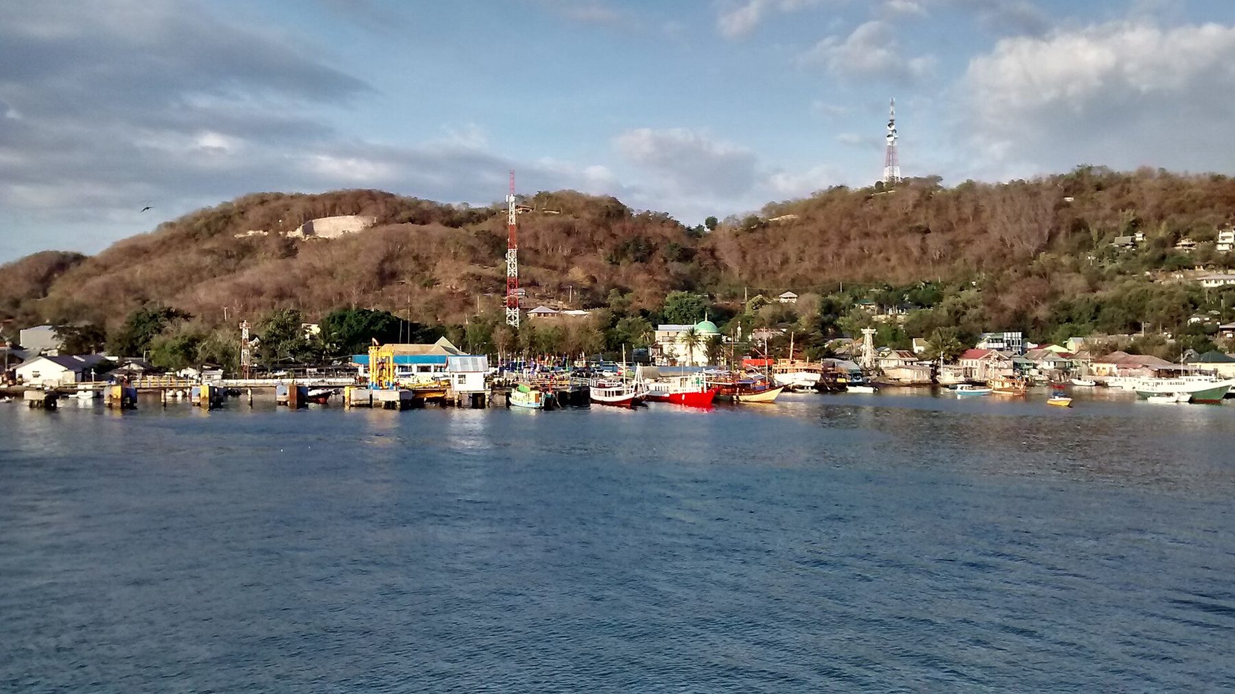 A wide paved street in Labuan Bajo lined with low shopfronts, parked motorbikes and hills rising behind the rooftops