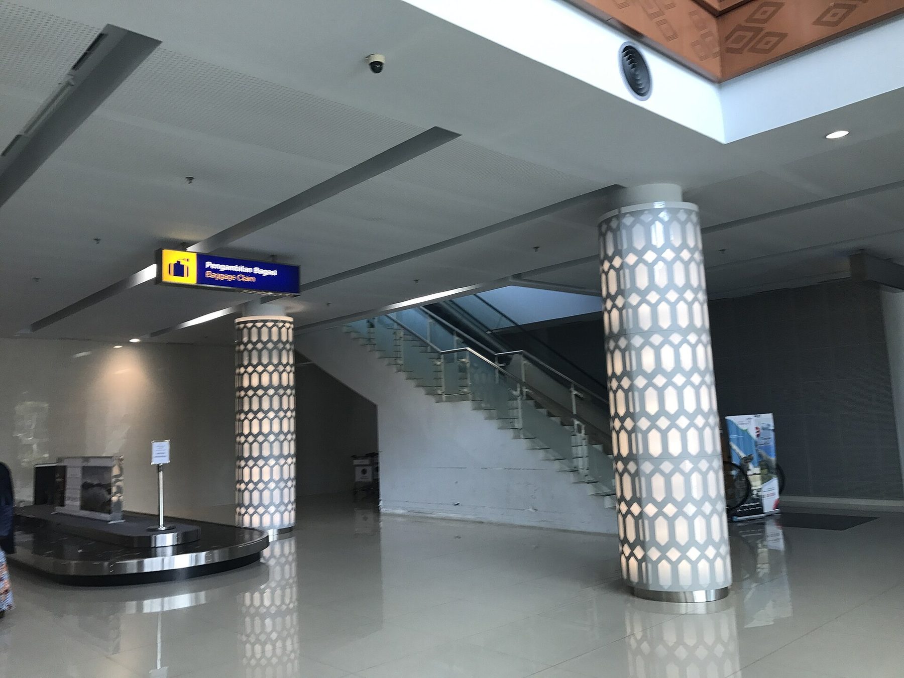 A single baggage carousel under a yellow and blue sign reading Pengambilan Bagasi, Baggage Claim, beside two patterned columns and an escalator in a quiet arrivals hall