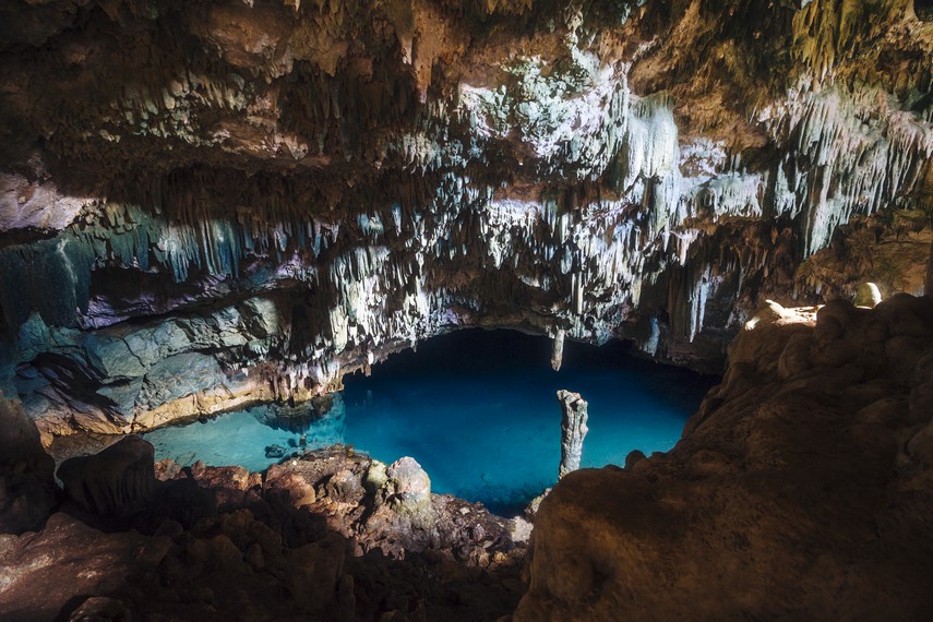 Sunlight falls through an opening in a limestone cave roof onto a still pool of water inside a rocky chamber