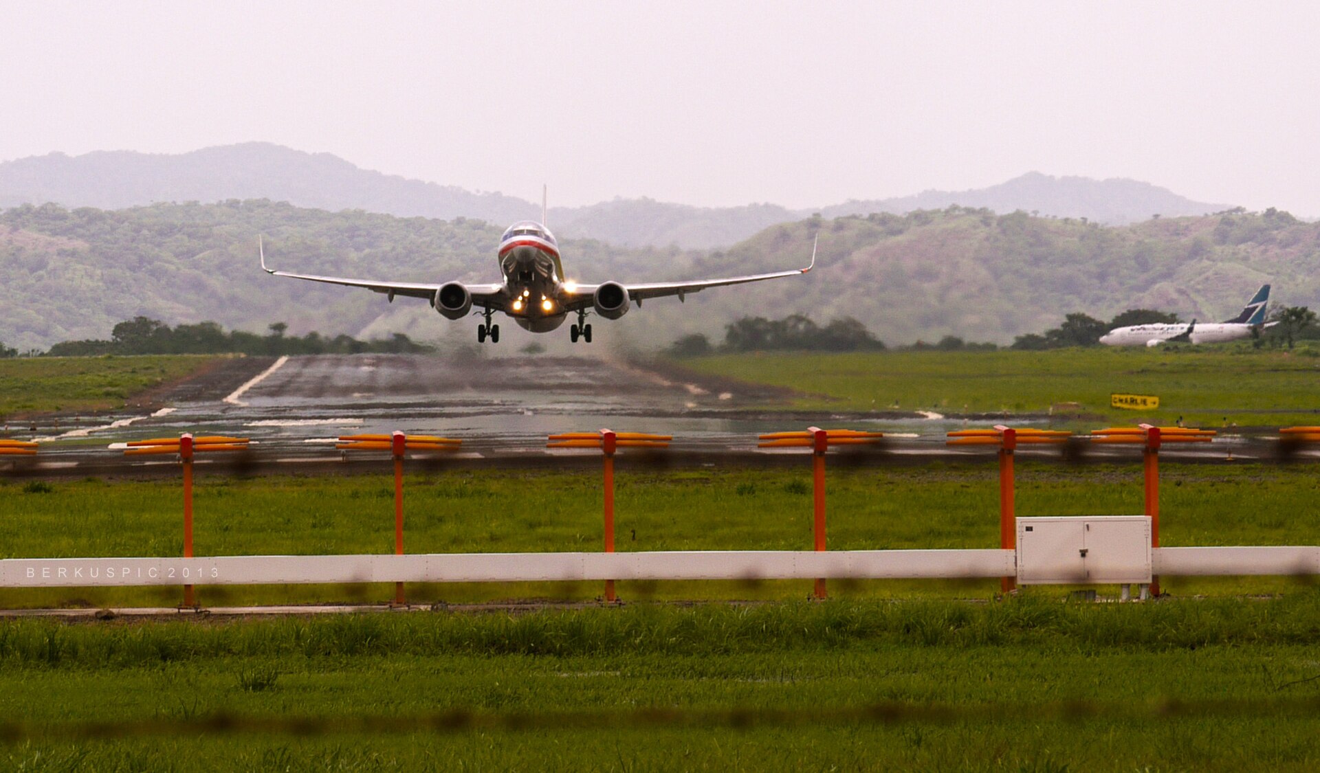 An American Airlines Boeing 737 lifting off head-on from a wet runway, green hills behind it and a WestJet jet waiting to the right